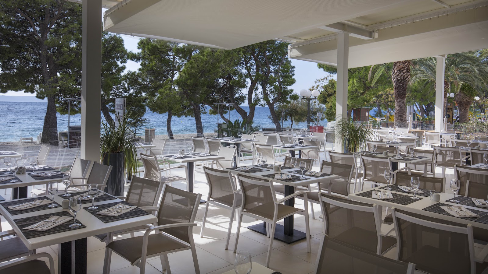 Covered beachfront dining area at Bluesun Hotel Alga with white tables, chairs, and ocean view framed by pine trees.