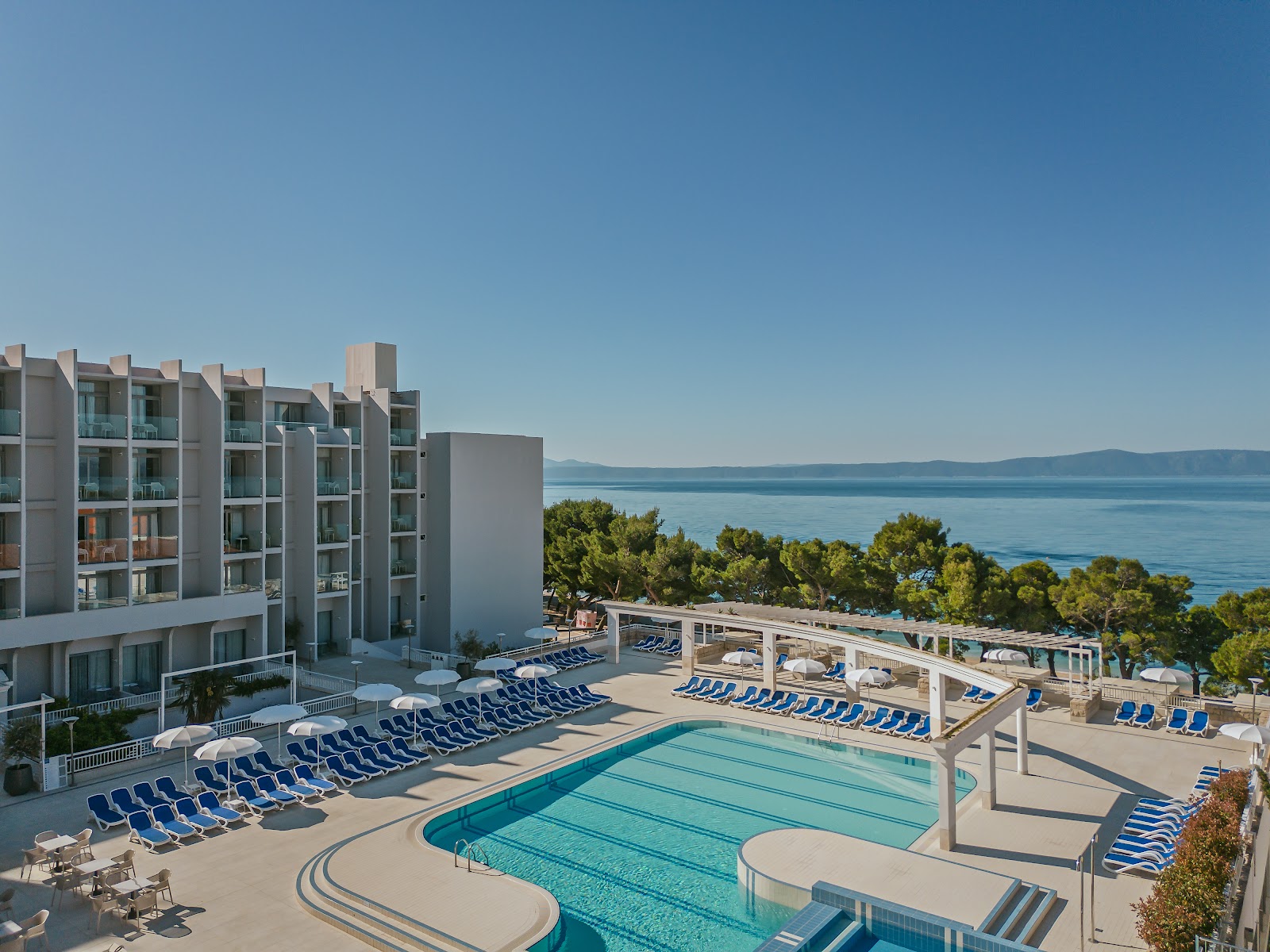 Two children wade in a shallow curved pool at Bluesun Hotel Alga, with a lap pool and loungers visible beyond under a clear sky.