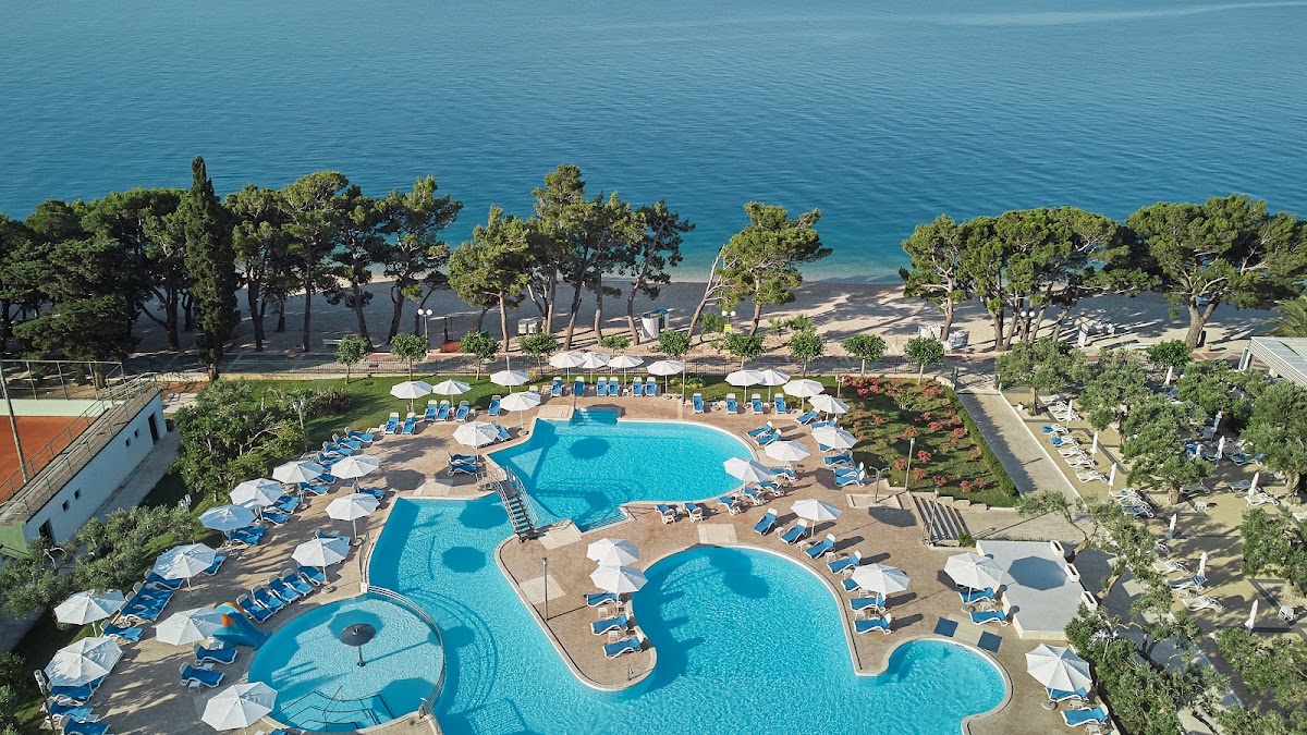 Multiple connected pools with blue loungers and white umbrellas, surrounded by pine trees, with a beach and turquoise sea beyond.