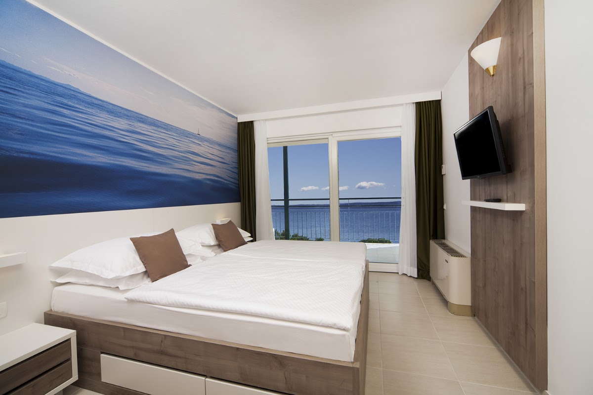Resort guestroom with double bed, ocean view balcony, and wall-mounted TV overlooking the Adriatic Sea.