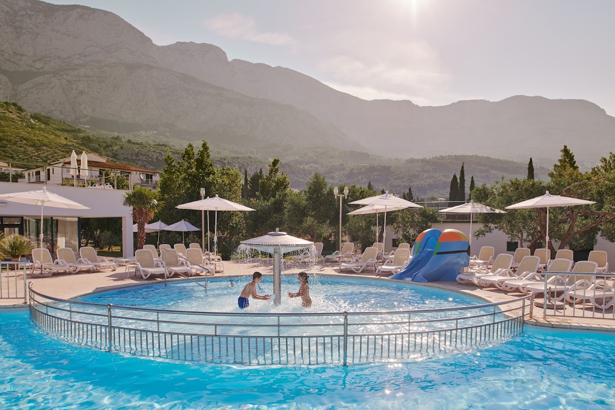 Resort pool with children playing in shallow fenced section, lounge chairs and umbrellas surrounding deck, mountains rising in background.