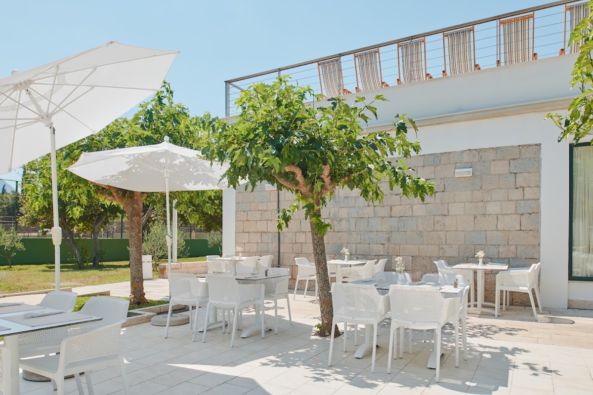 White umbrellas shade dining tables on a stone terrace at Bluesun Hotel Neptun, with mature trees and a modern building facade visible.