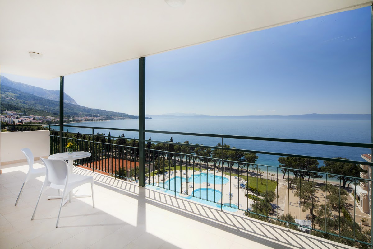 Balcony with white chairs overlooking a pool, beach, and calm blue sea with forested coastline and mountains beyond.