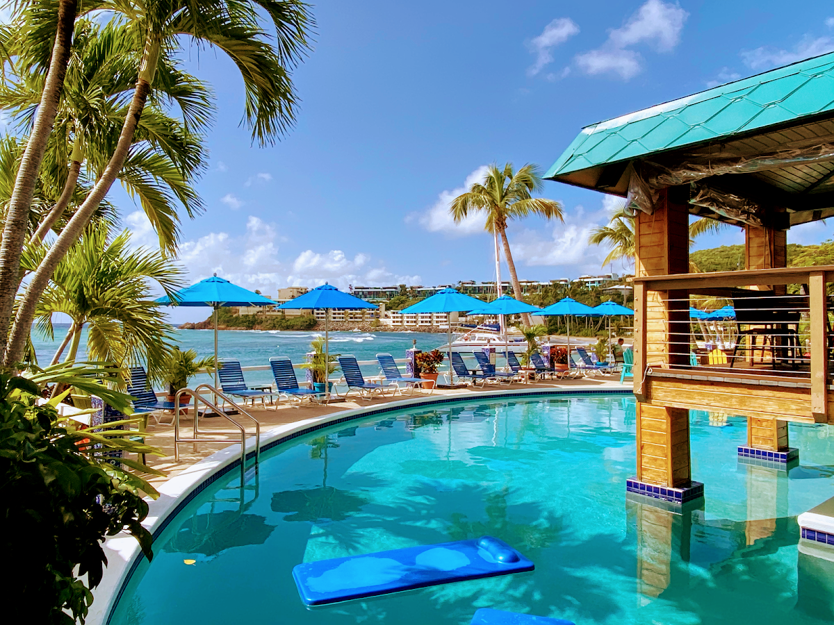 Curved turquoise pool with wooden bar structure and blue umbrellas, overlooking beach and calm Caribbean waters at Bolongo Bay Beach Resort.