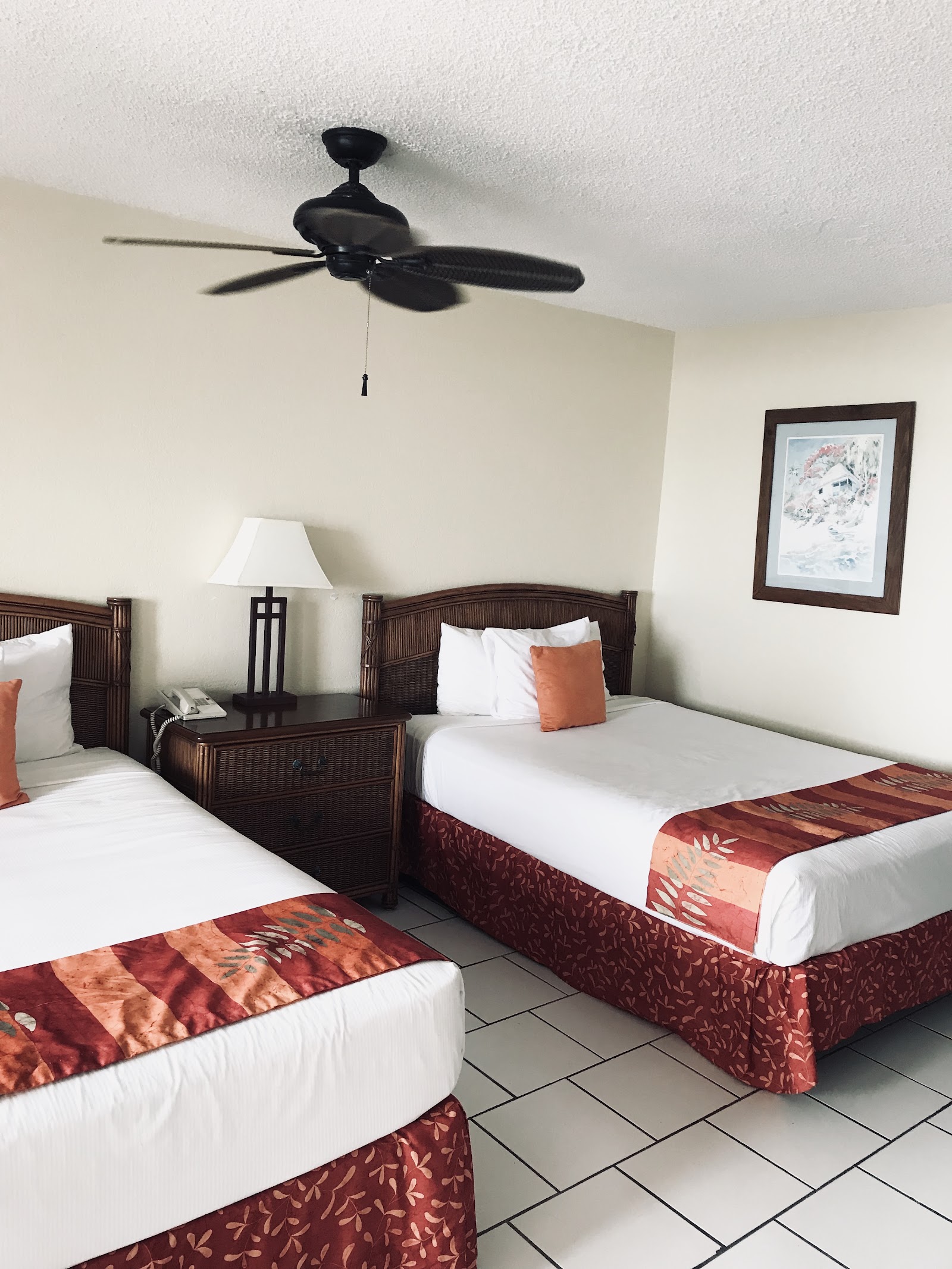 Hotel room with two double beds with rust-colored runners, dark wood furniture, white walls, ceiling fan, and framed artwork.