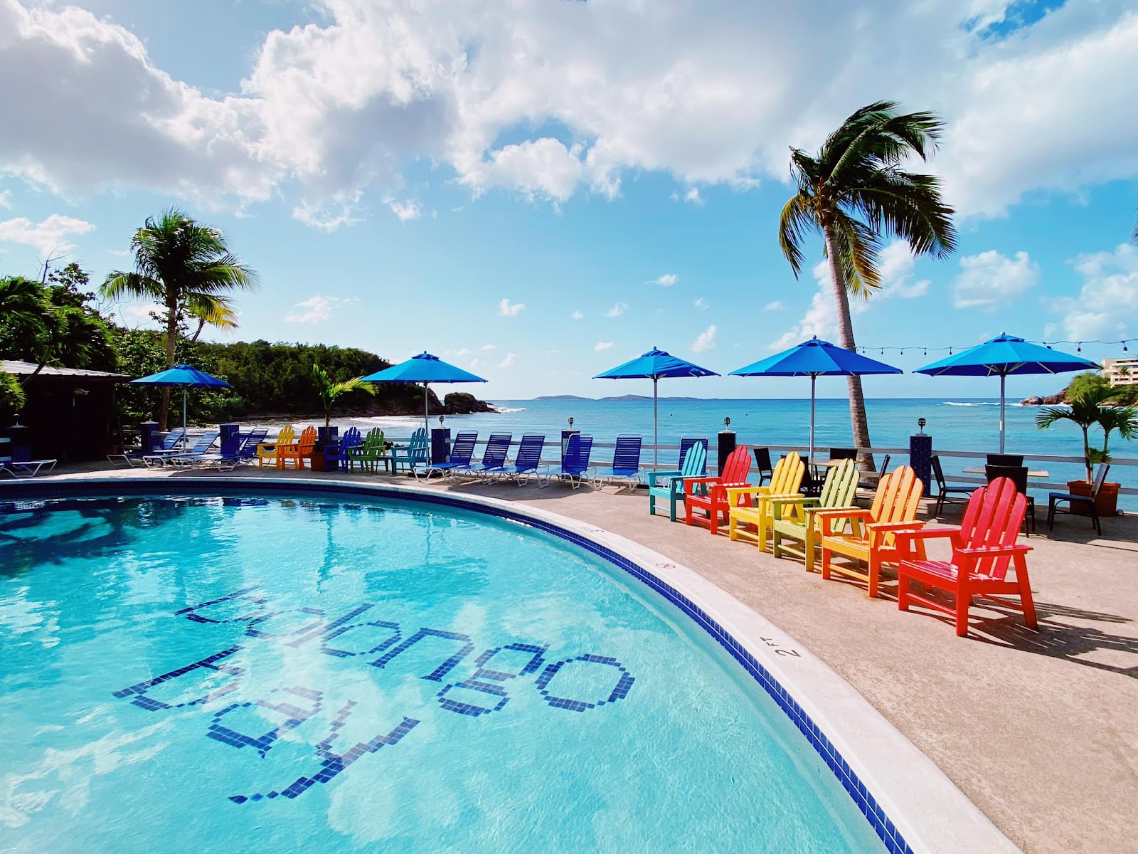 Bolongo Bay Beach Resort's pool and beach area with colorful Adirondack chairs, blue umbrellas, palm trees, and turquoise Caribbean water under a partly cloudy sky.
