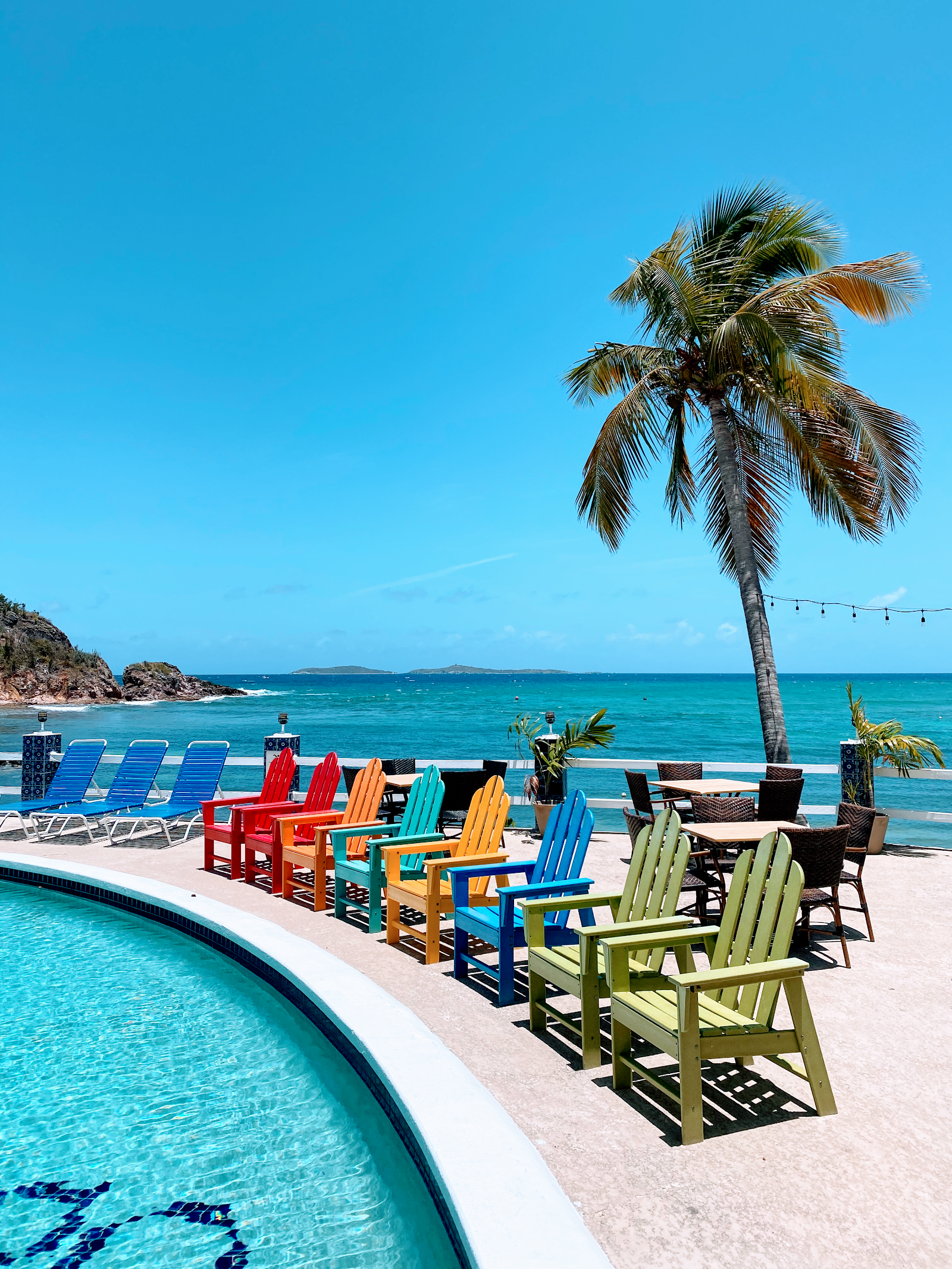 Colorful Adirondack chairs line a sandy beach at Bolongo Bay Beach Resort, with a pool in the foreground and turquoise Caribbean Sea beyond.