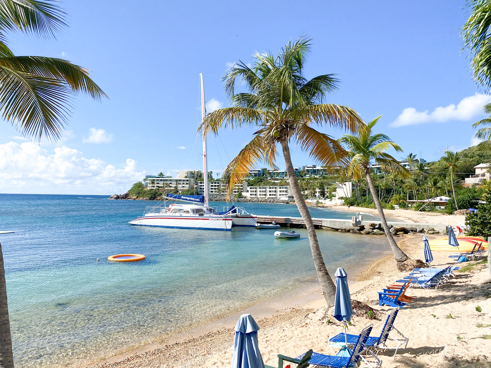 Beach cove at Bolongo Bay with sailboat at dock, palm trees, blue loungers, and hillside resort buildings under clear sky.