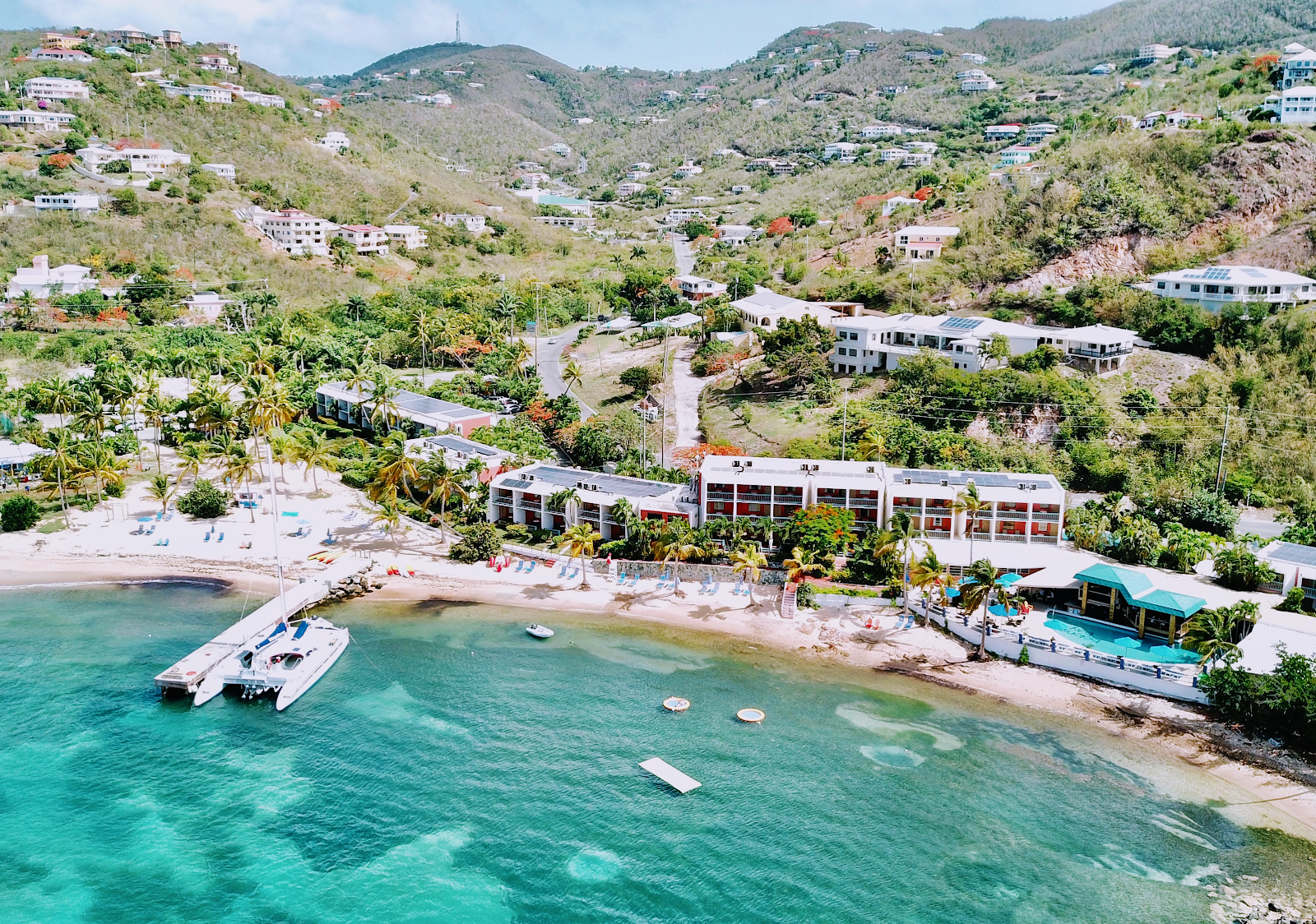 Bolongo Bay Beach Resort's beachfront with turquoise water, moored boats, sandy shore, and modern white buildings nestled against forested hillside.