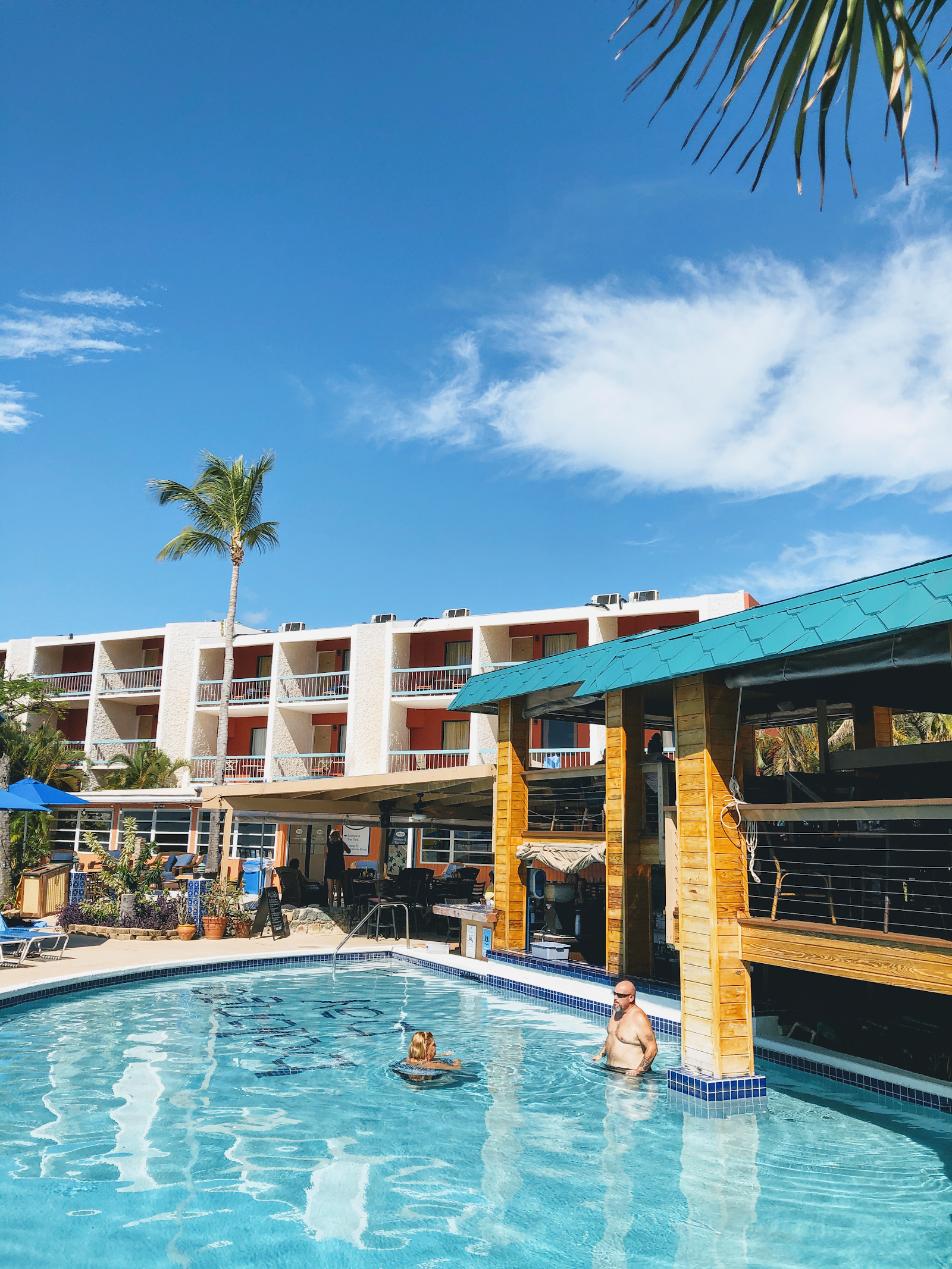Two guests swim in the turquoise pool at Bolongo Bay Beach Resort with a wooden bar pavilion and multi-story white building behind them under clear blue sky.