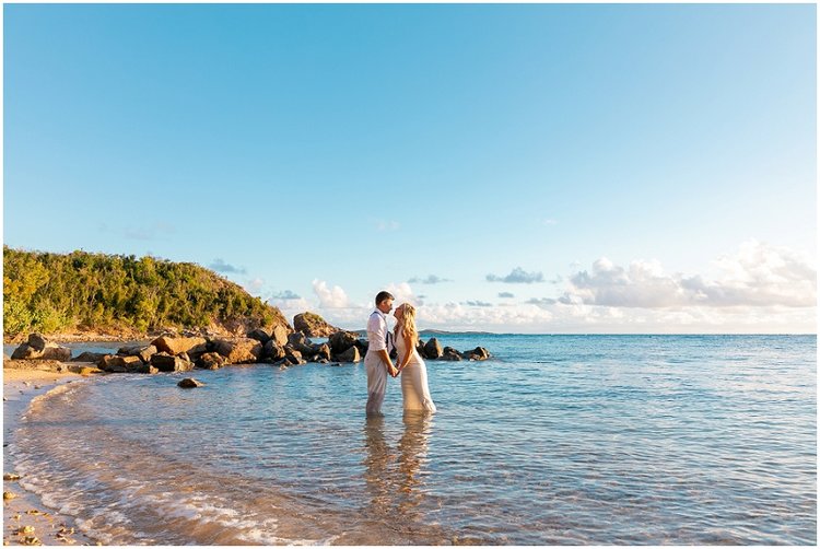 Couple wading in clear turquoise water at Bolongo Bay Beach Resort with rocky shoreline and tropical vegetation.