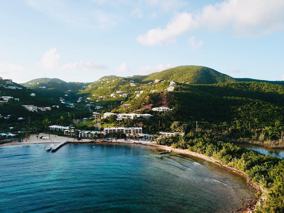 Bolongo Bay Beach Resort sits on a sandy beach along turquoise water, with lush green hills and residential buildings scattered across the hillside behind it.