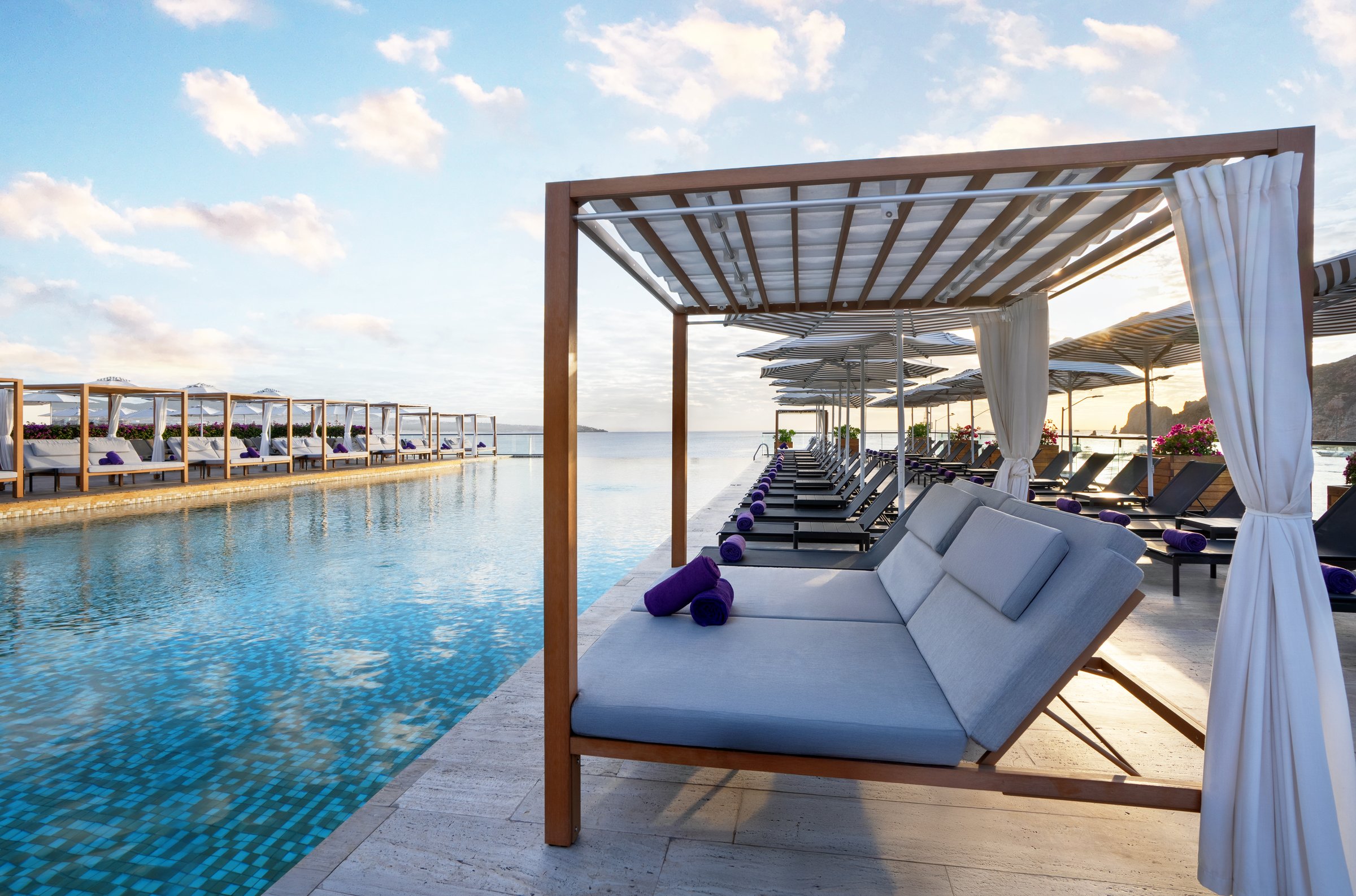 Wooden poolside cabana with gray cushions and purple towels beside the blue beach club pool.
