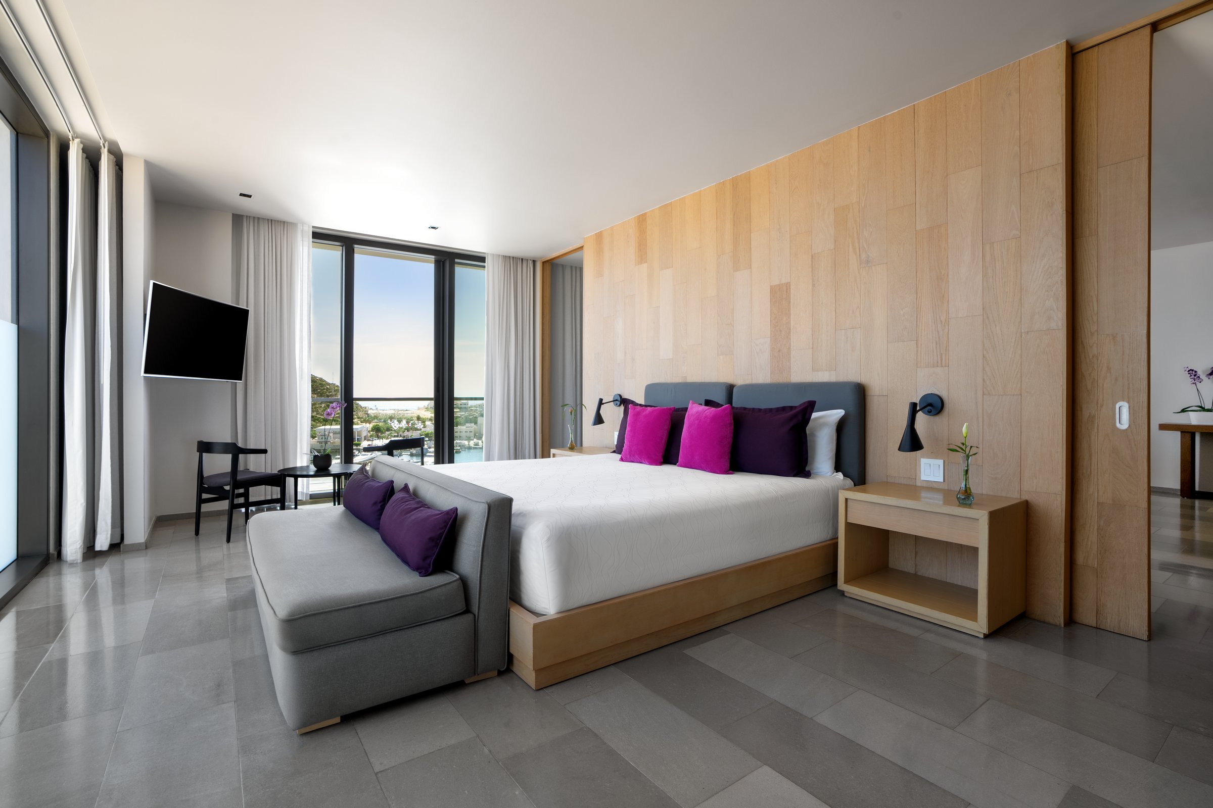 Xhale Club master suite with a king bed, magenta accent pillows, light wood paneling and gray tile floors.