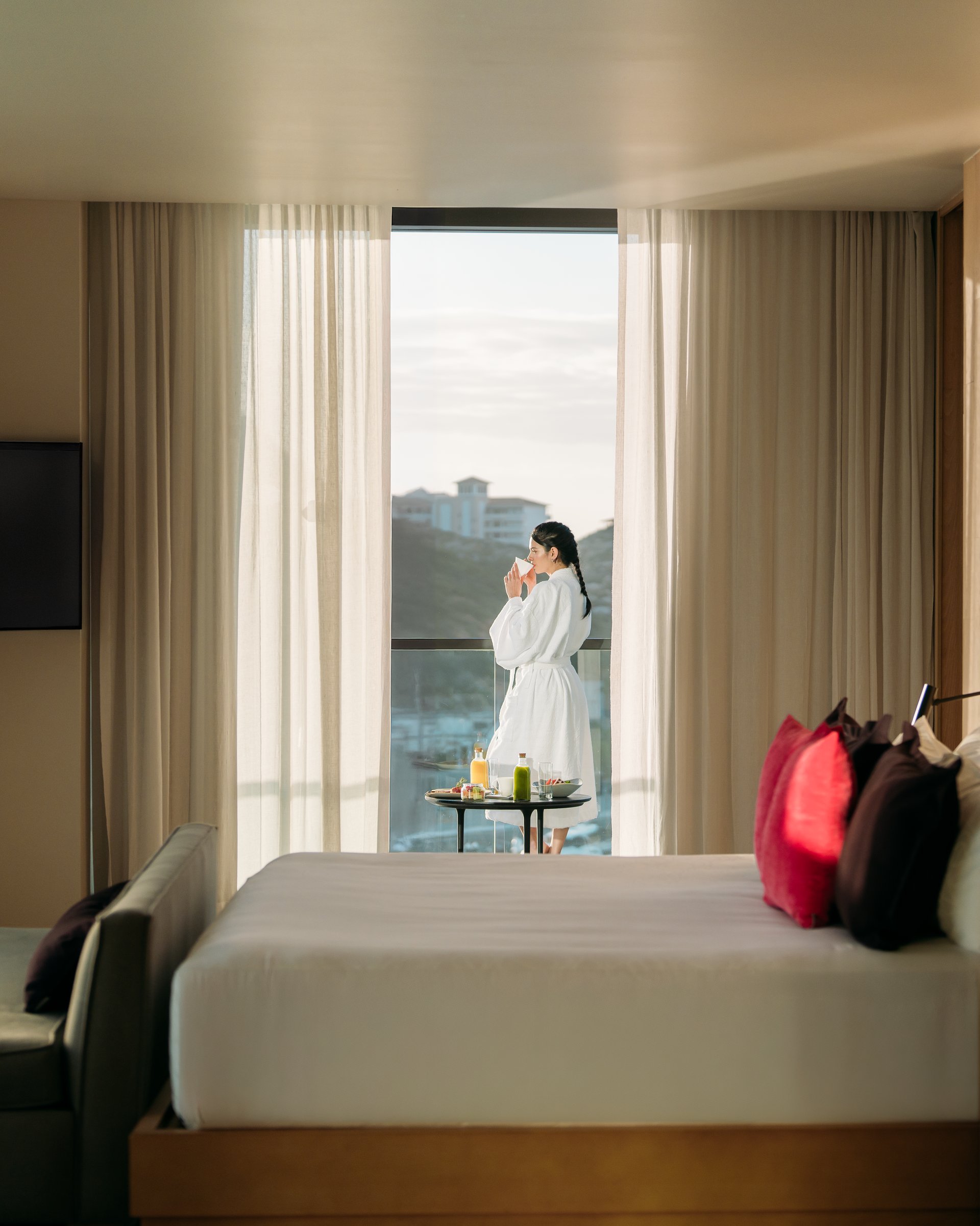 Suite interior with the corner of a bed and a floor-to-ceiling window to a balcony and hills.