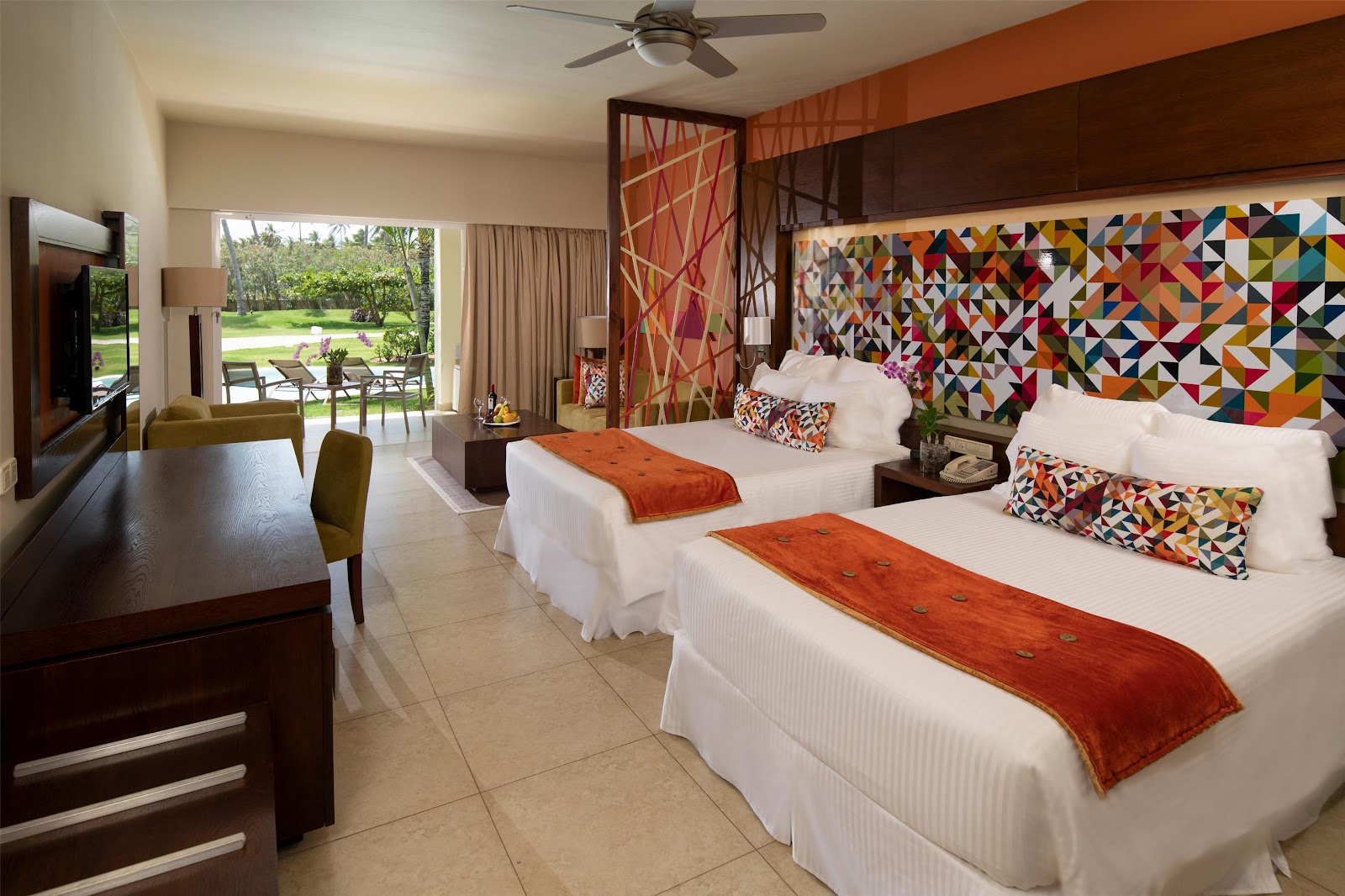 Resort room with two queen beds dressed in white linens and orange throws, colorful geometric headboard, and sliding glass doors opening to a lawn and pool area.