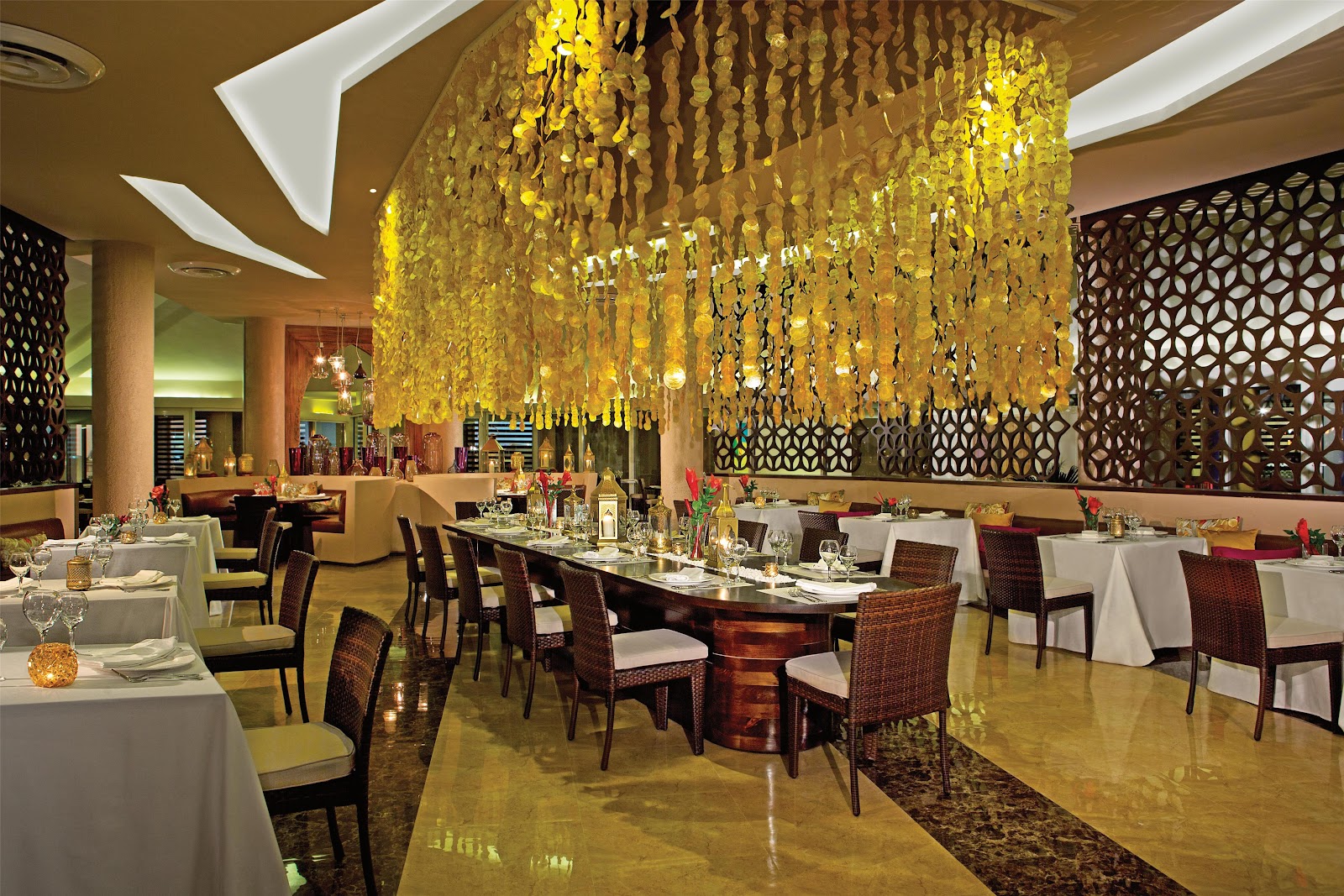 Fine dining restaurant with hanging yellow floral installations, white-clothed tables, woven chairs, and decorative geometric screens throughout the space.