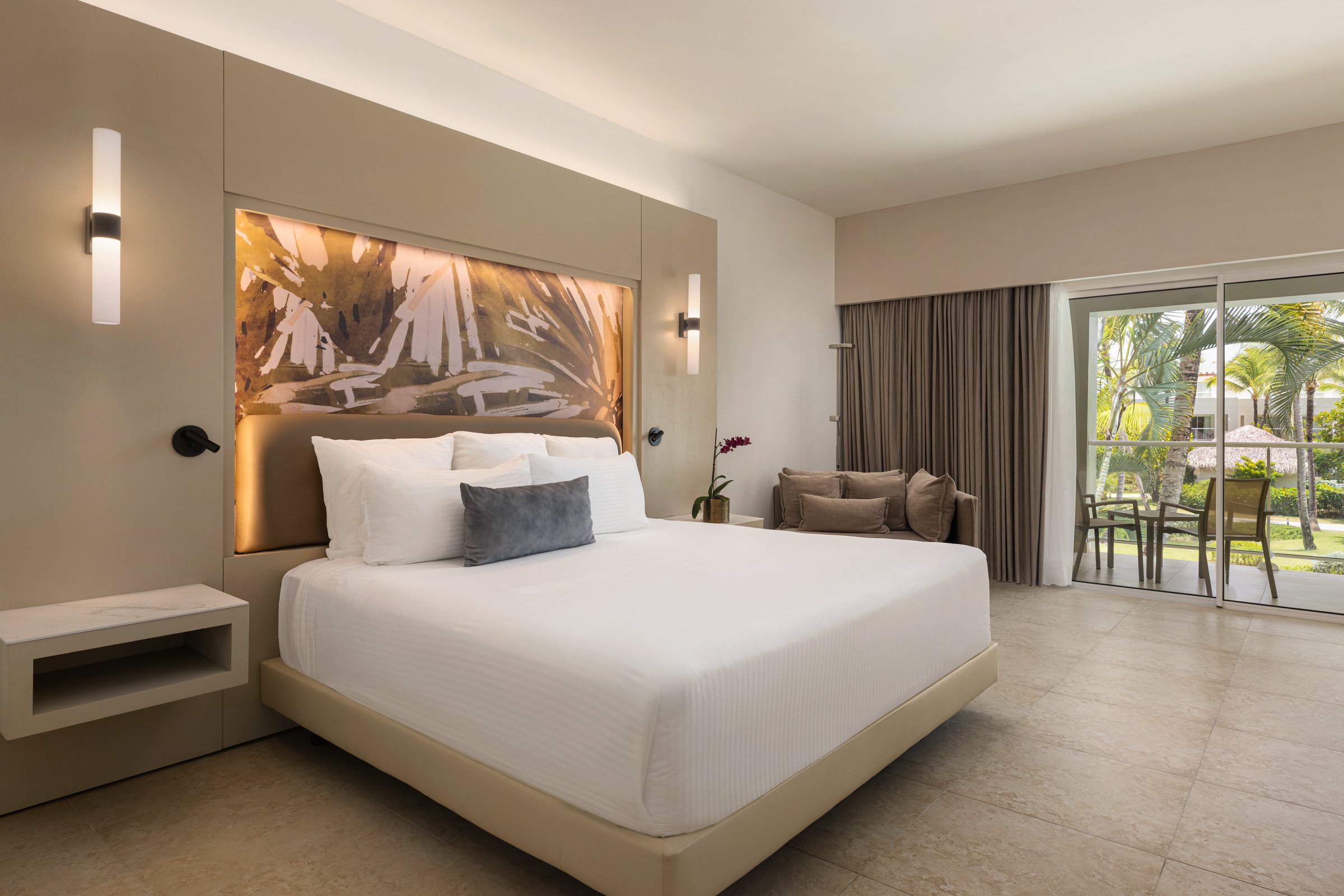 Allure junior suite with a king bed against a lit gold palm-leaf panel and glass doors to a garden-view balcony.