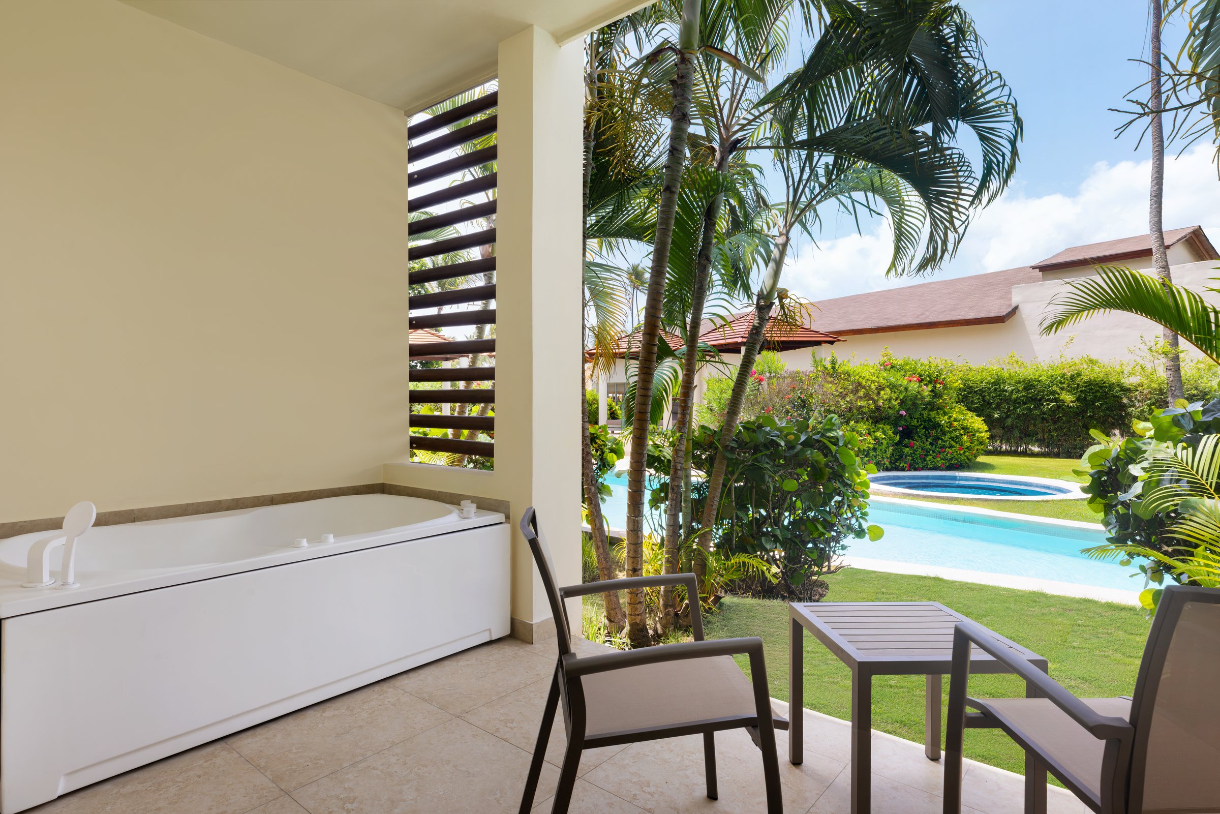 Suite terrace with a white whirlpool tub and two black chairs over a green lawn, palms and a blue pool.