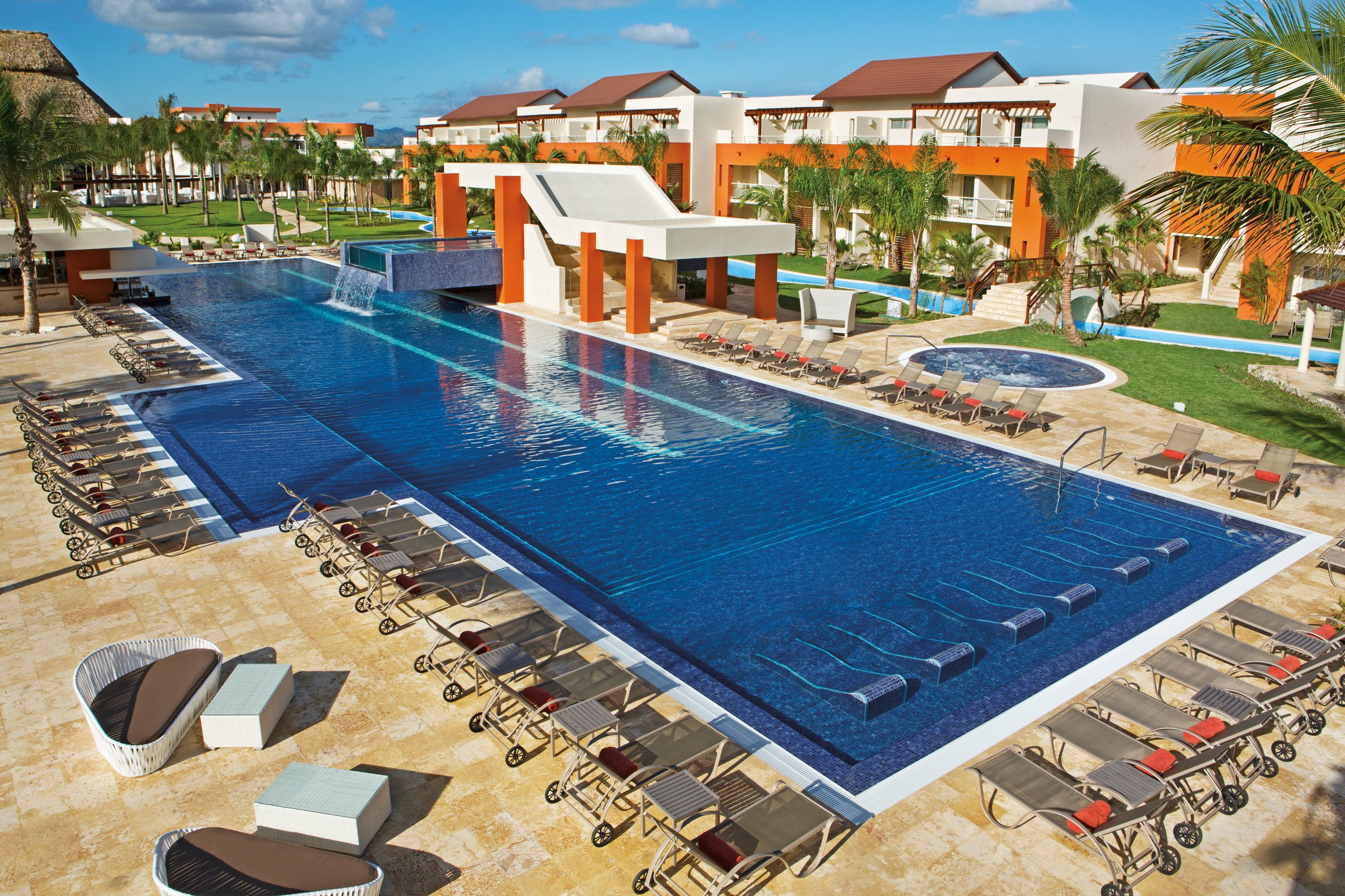 High view of the long rectangular pool with grey loungers, a waterfall platform, submerged lounge chairs and orange and white buildings.
