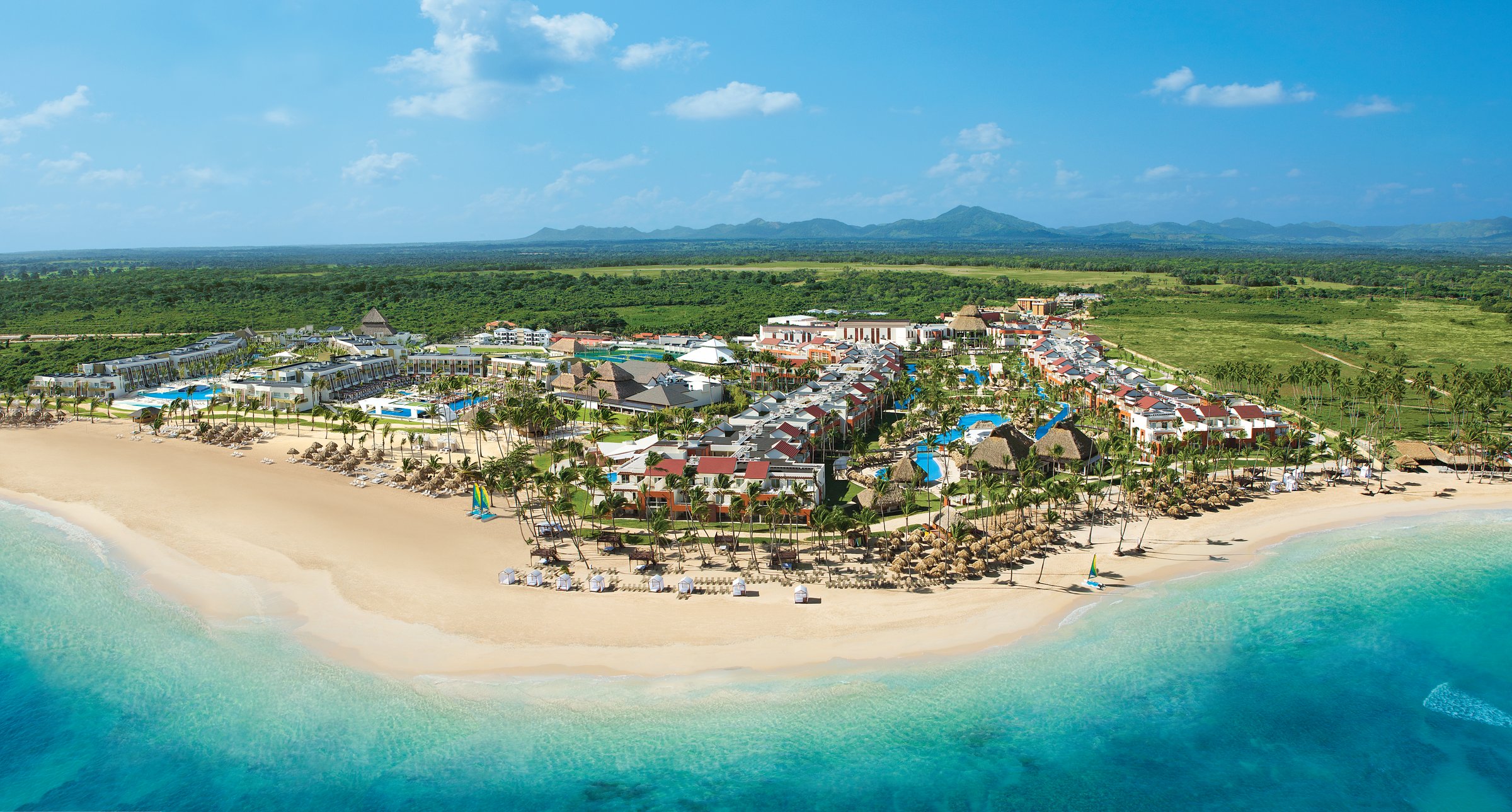 Aerial view of the white resort buildings between dense tropical forest and a white beach dotted with palapas.