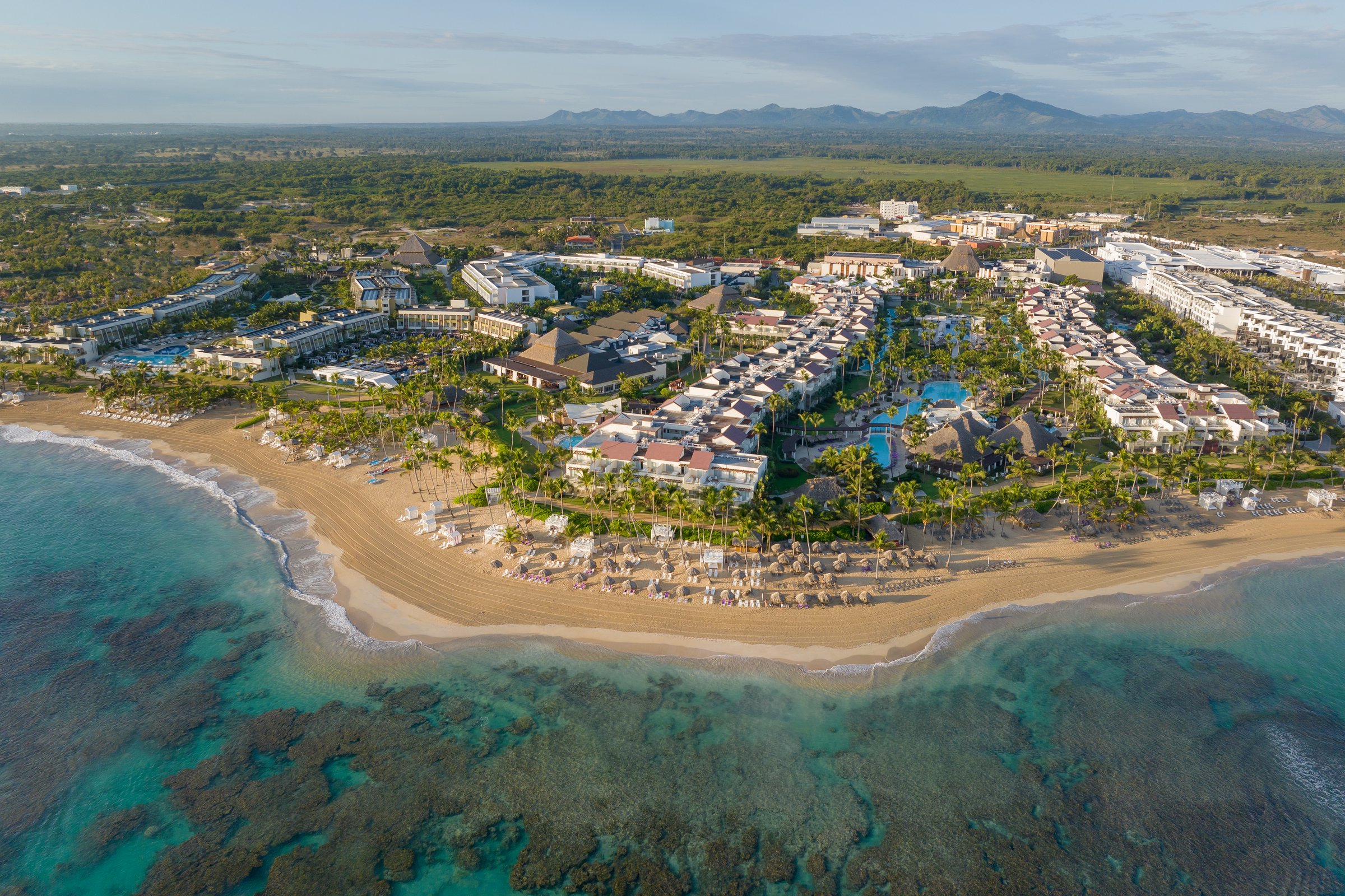 High aerial view of the whole resort on its point of land with pools, green grounds, the offshore reef and distant mountains.