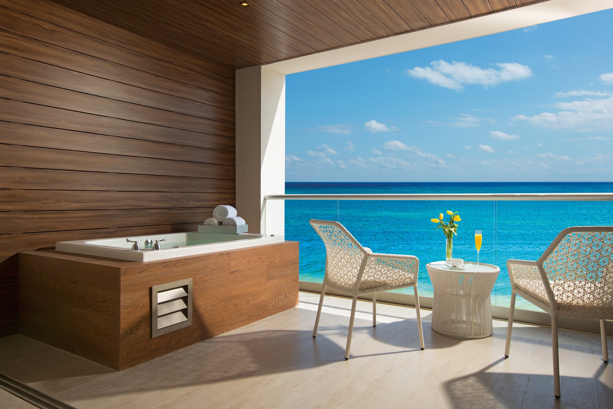 Private terrace with a wooden-paneled whirlpool tub, two chairs and a glass railing over the turquoise ocean.