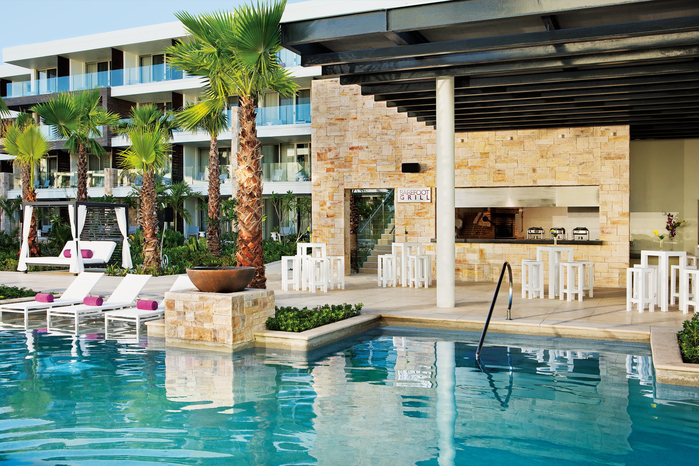 Barefoot Grill beside the turquoise pool, with white loungers, stone planters and palm trees in front of the resort buildings.