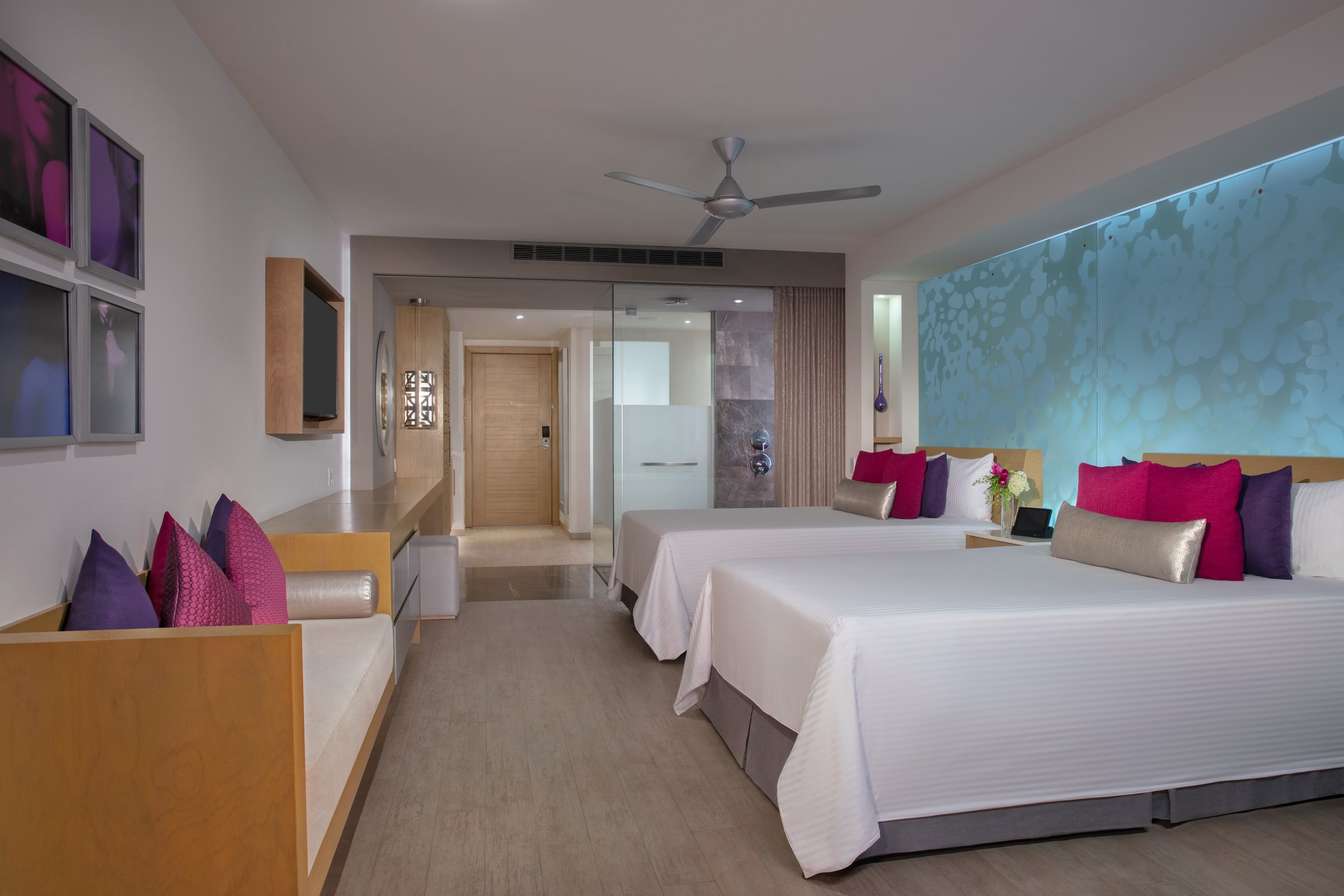 Junior suite with two double beds, purple accents, a wall TV on a wooden console and a ceiling fan.