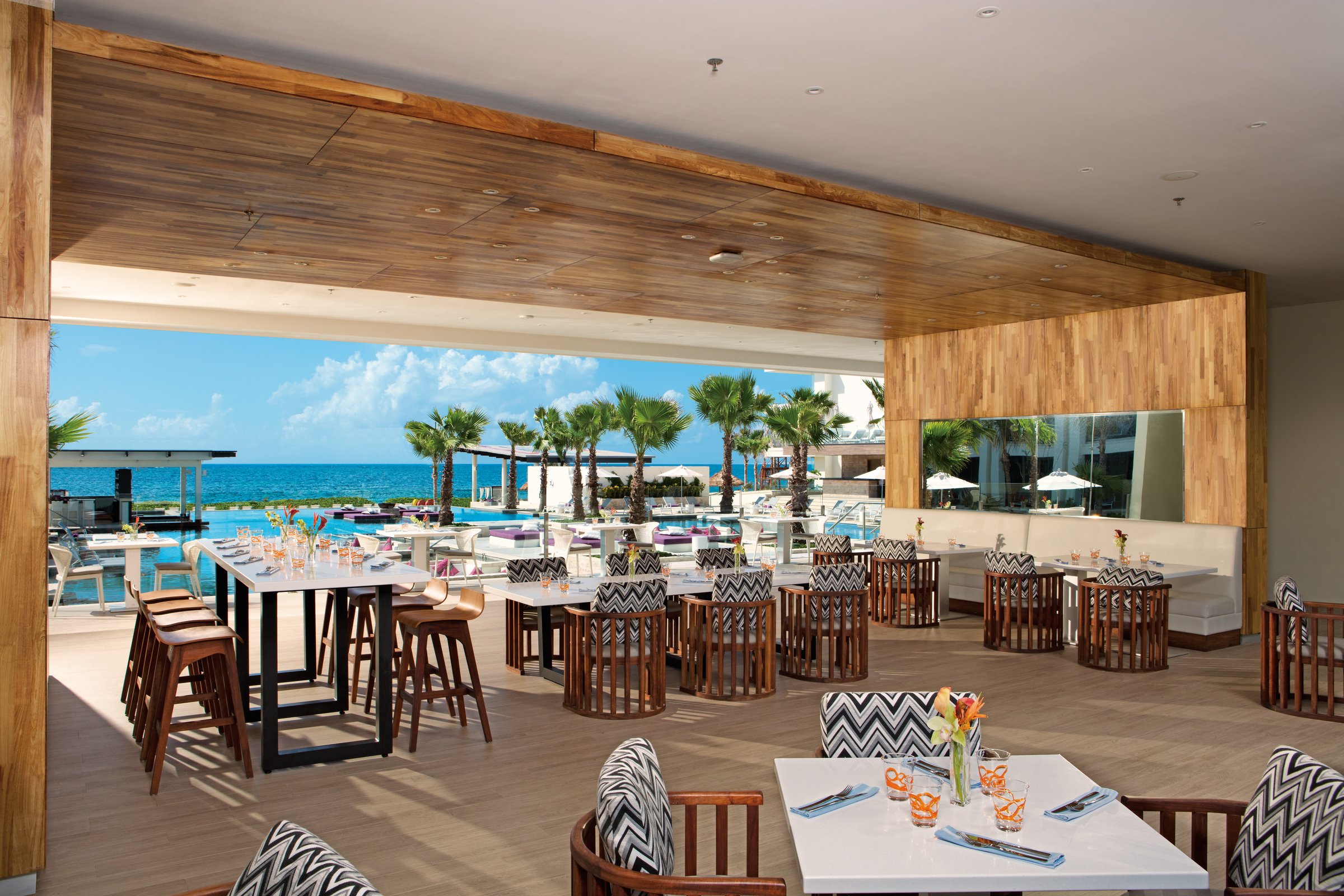 Open-air Bites restaurant with a light wood ceiling and tables set with white cloths and orange accents, looking onto the pool deck and sea.