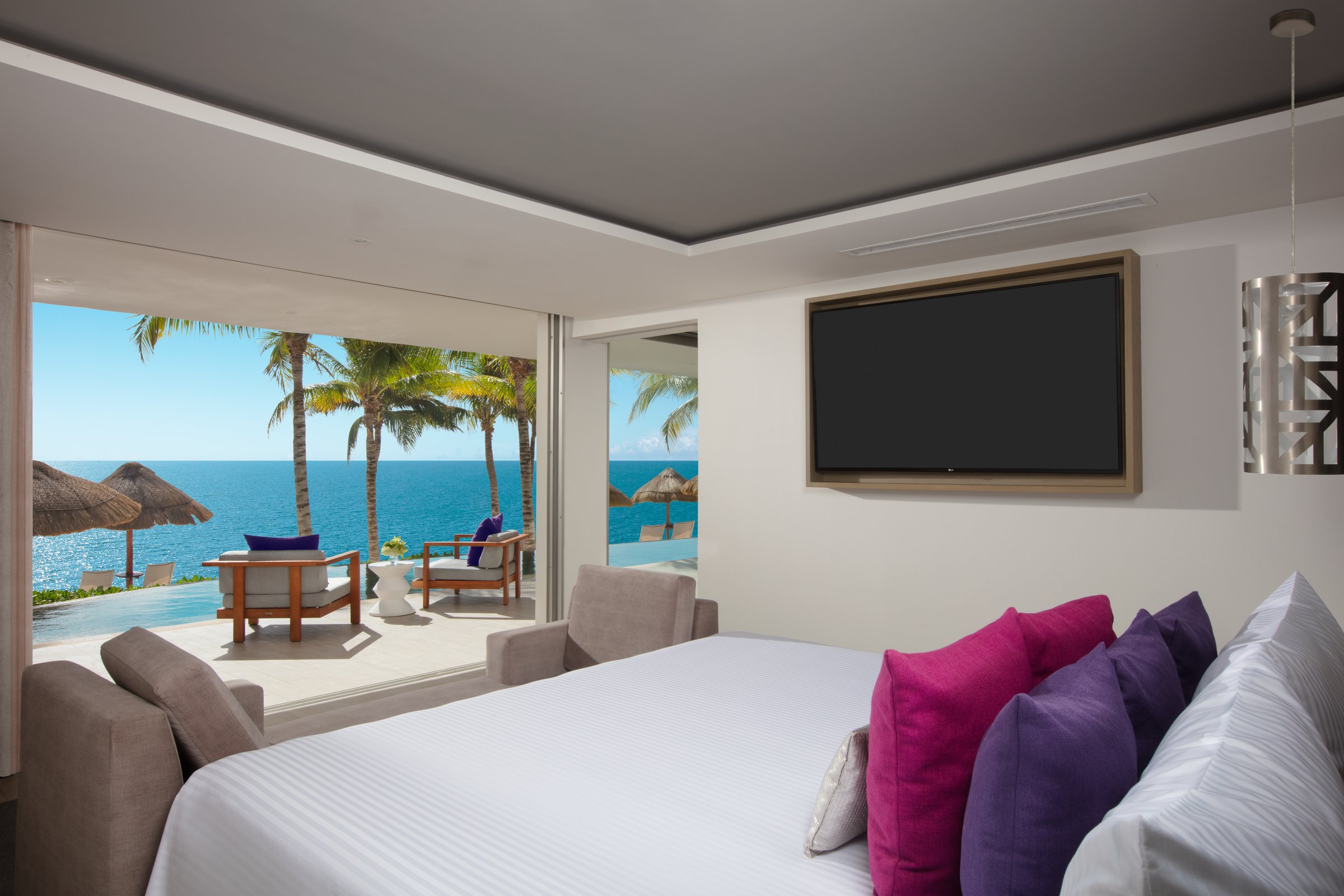 Presidential swim-out suite bedroom with a large bed and floor-to-ceiling glass doors to a balcony, palms and the bright blue ocean.