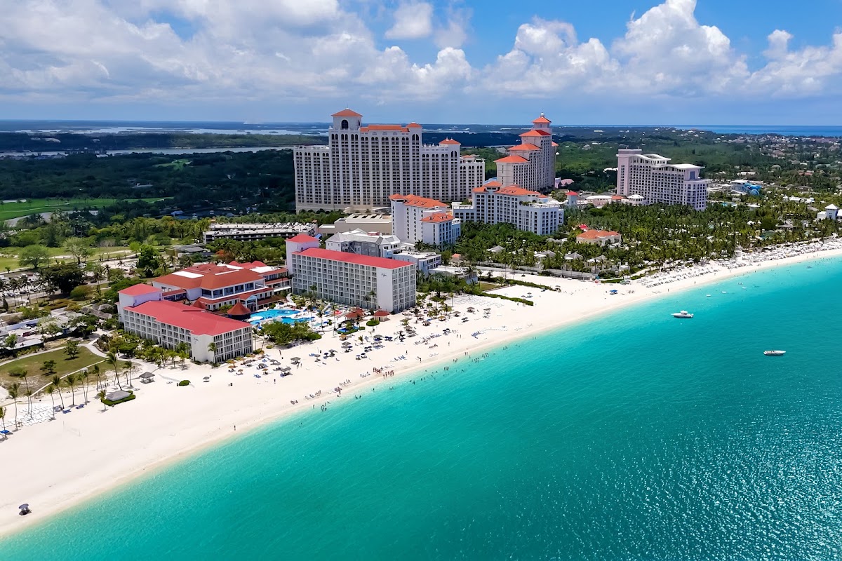 Aerial view of Breezes Resort Bahamas with pink-roofed buildings, white sand beach, turquoise water, and palm trees lining the oceanfront.