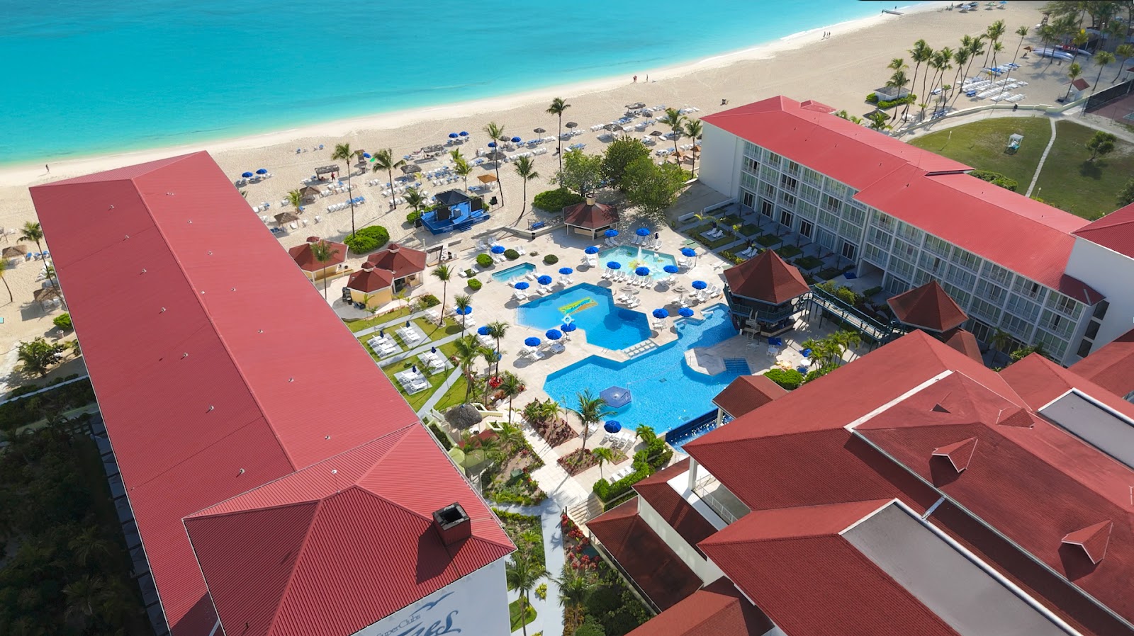 Aerial view of Breezes Resort Bahamas with red-roofed buildings, blue pool with umbrellas, and pristine beach with palm trees beyond.
