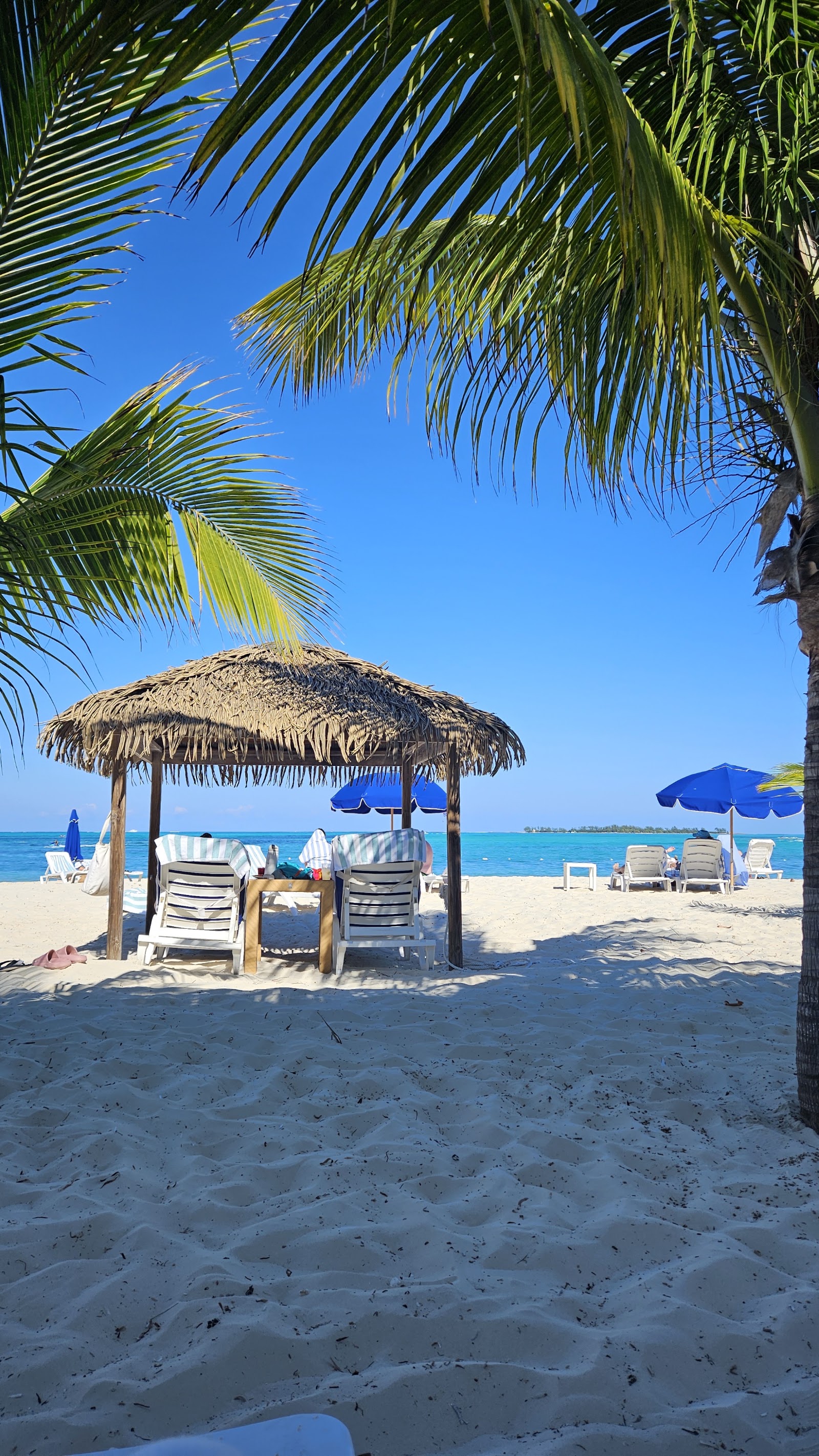 Palm trees and beach vegetation frame the white sand beach at Breezes Resort Bahamas, with thatched umbrellas, lounge chairs, and turquoise water beyond.