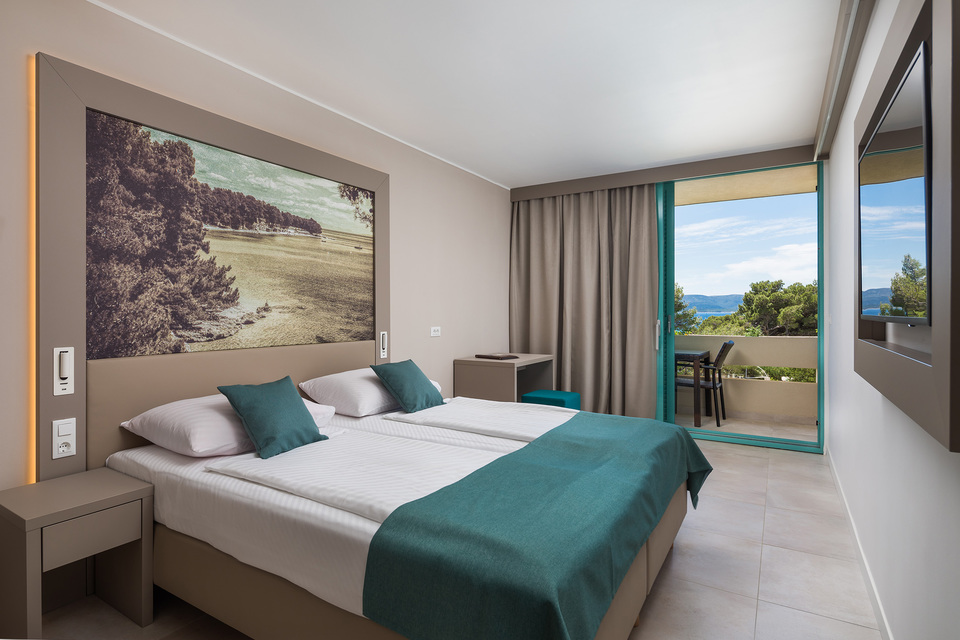 Modern bedroom with double bed, teal accents, and sea view through glass doors at BRETANIDE Sport & Wellness Resort.