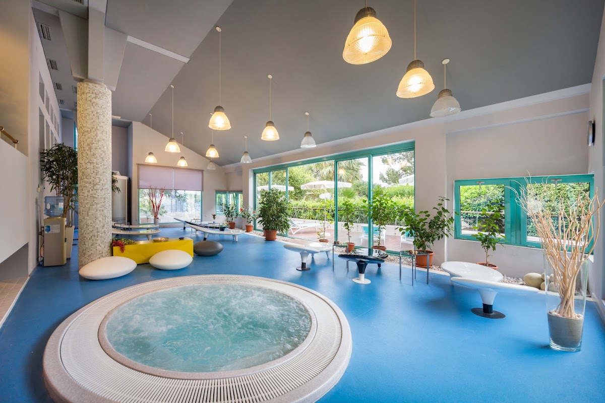 Indoor spa area with round hot tub, blue flooring, pendant lights, seating pods, and views of tropical grounds through large windows.
