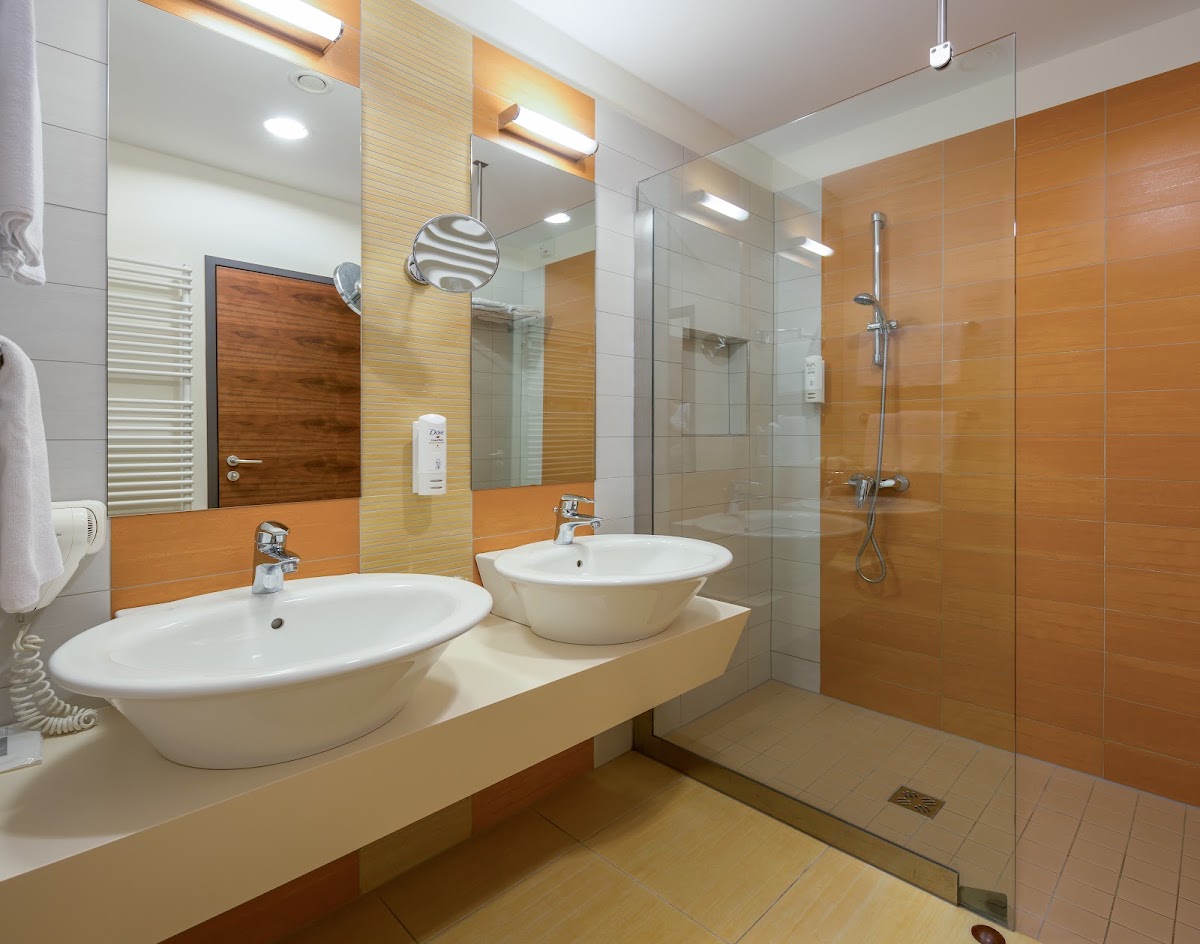 Modern bathroom with two white bowl sinks, orange tile accents, large mirror, and walk-in shower with glass enclosure at BRETANIDE Sport & Wellness Resort.