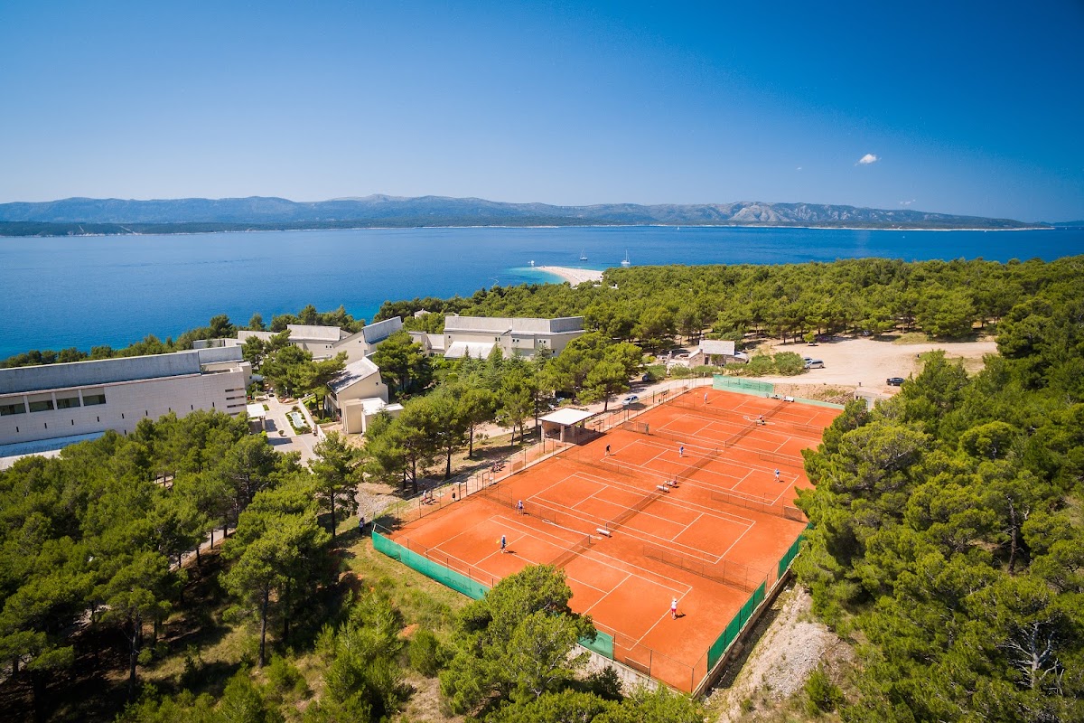 Aerial view of orange clay tennis courts surrounded by pine trees, with resort buildings and a blue Adriatic Sea coastline with mountains beyond.
