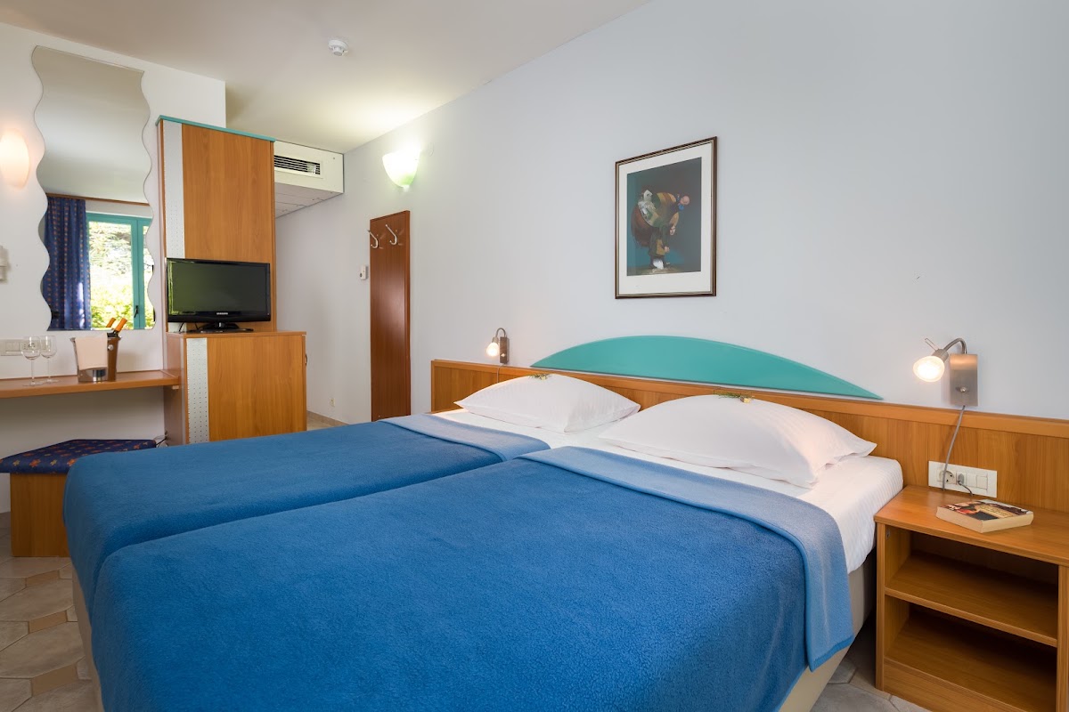 Hotel room with two beds dressed in blue and white linens, wooden furniture, TV, desk area, and turquoise headboard accent.