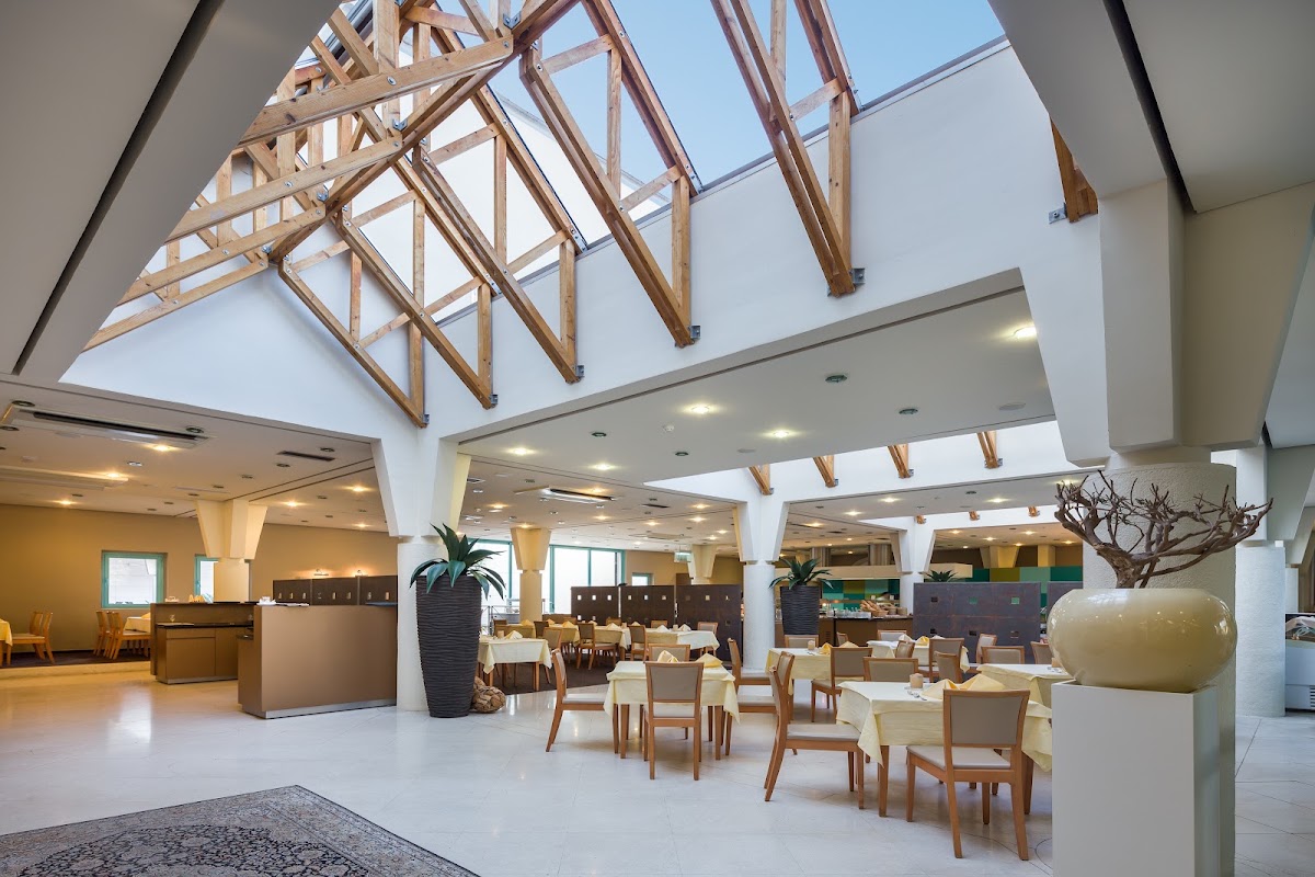 Spacious dining hall with wooden beam skylight, white walls, dining tables with cushioned chairs, potted plants, and modern pendant lighting.