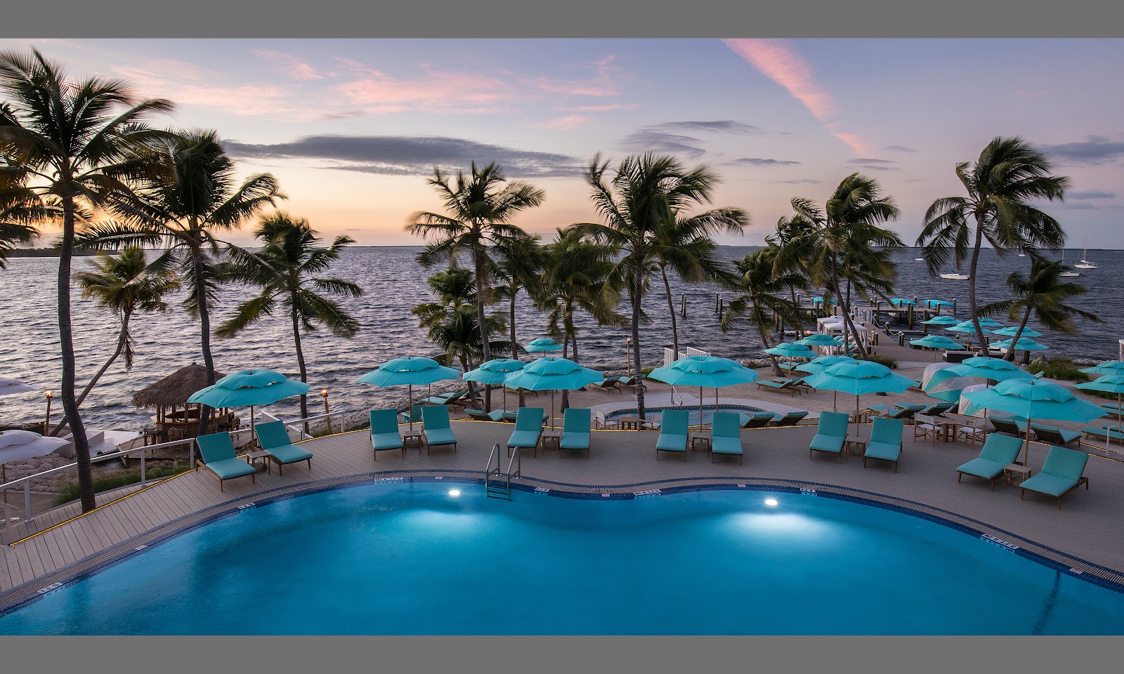 Illuminated pool at Bungalows Key Largo at dusk, with turquoise loungers and umbrellas overlooking the ocean and palm trees.
