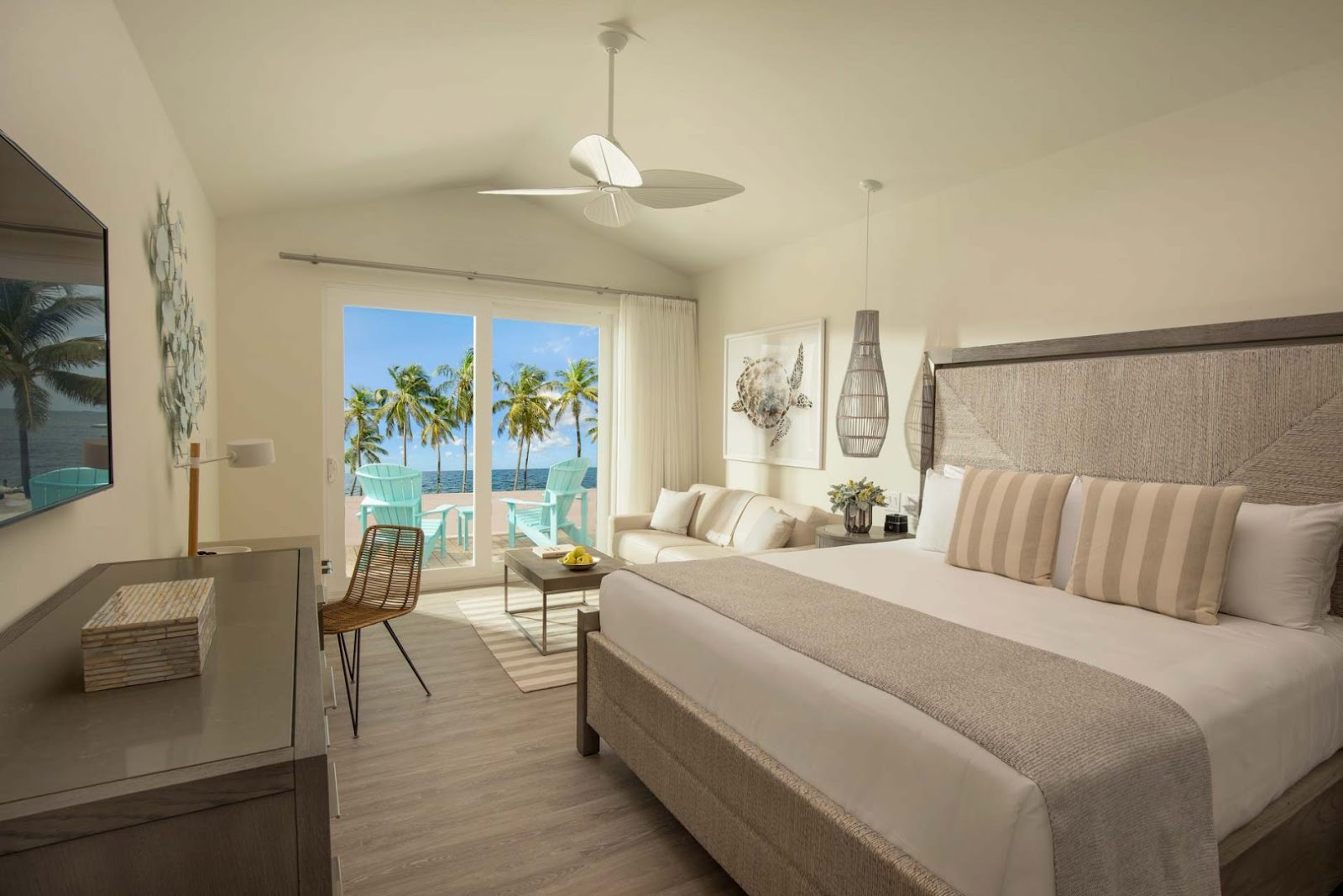 Beachfront guestroom at Bungalows Key Largo with king bed, ocean view balcony, palm trees, and turquoise lounge chairs on the sand.
