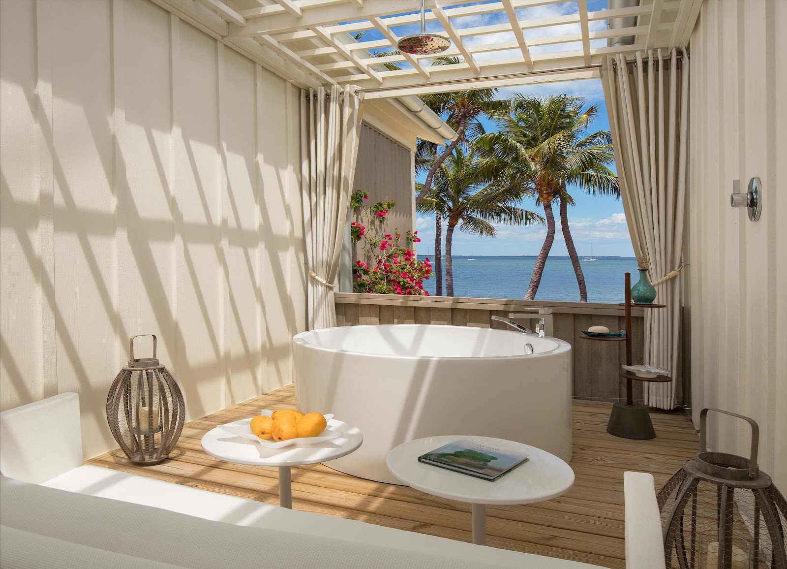 Luxurious bathroom with a freestanding white soaking tub, skylight ceiling, and ocean view framed by palm trees and pink flowers beyond the window.