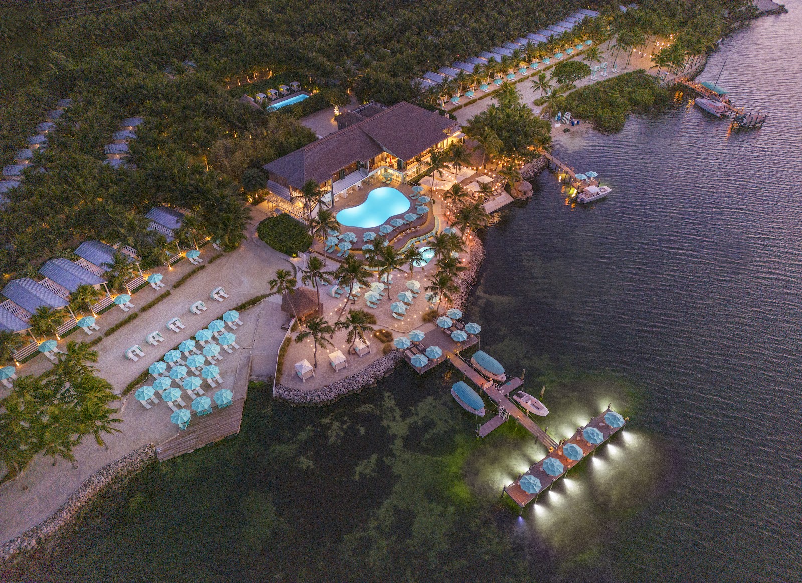 Aerial dusk view of Bungalows Key Largo with illuminated pool, beach loungers, main lodge, and lit dock extending into the water surrounded by mangroves.