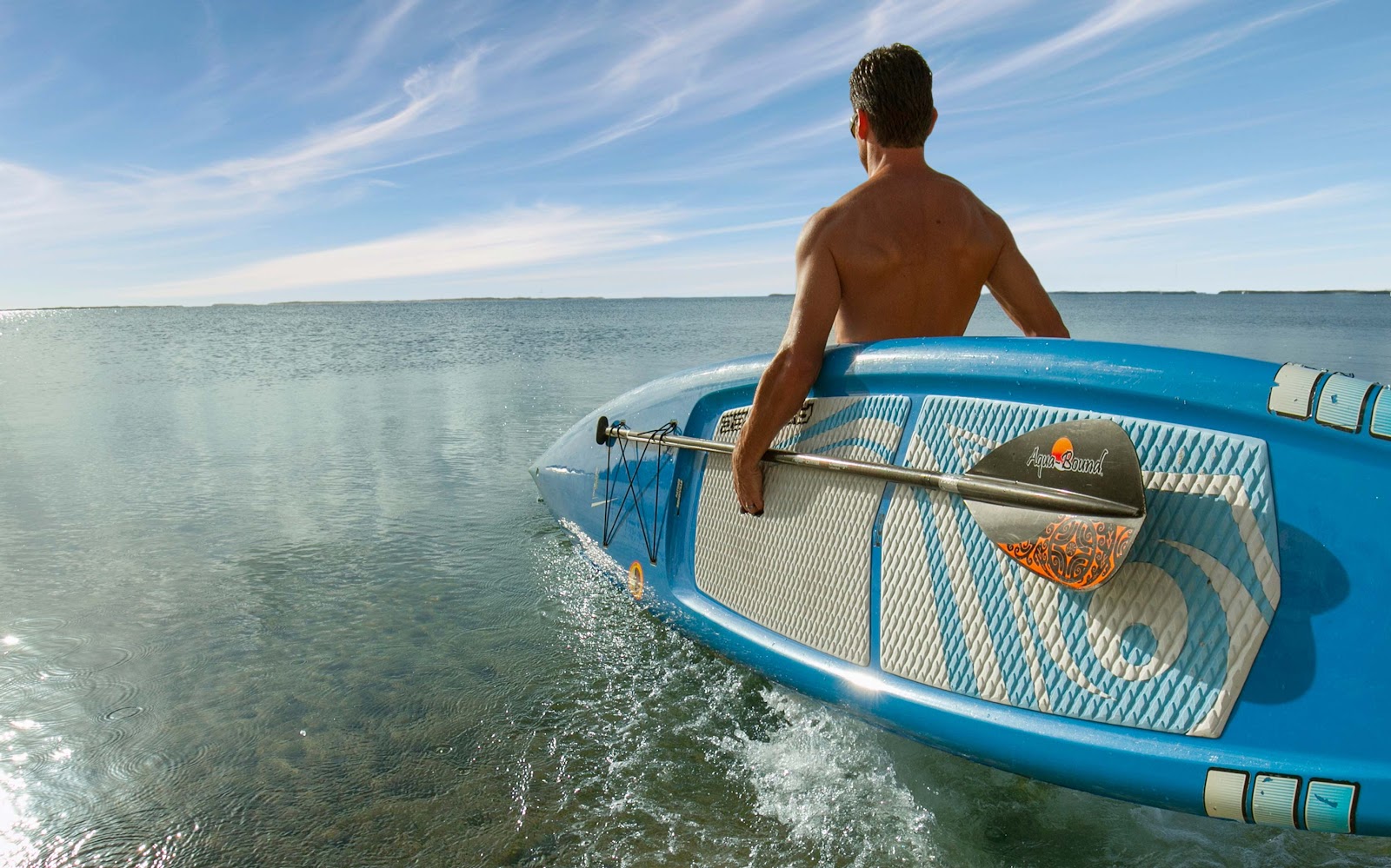 Man carrying paddle boards a blue inflatable board in shallow turquoise water under clear sky.