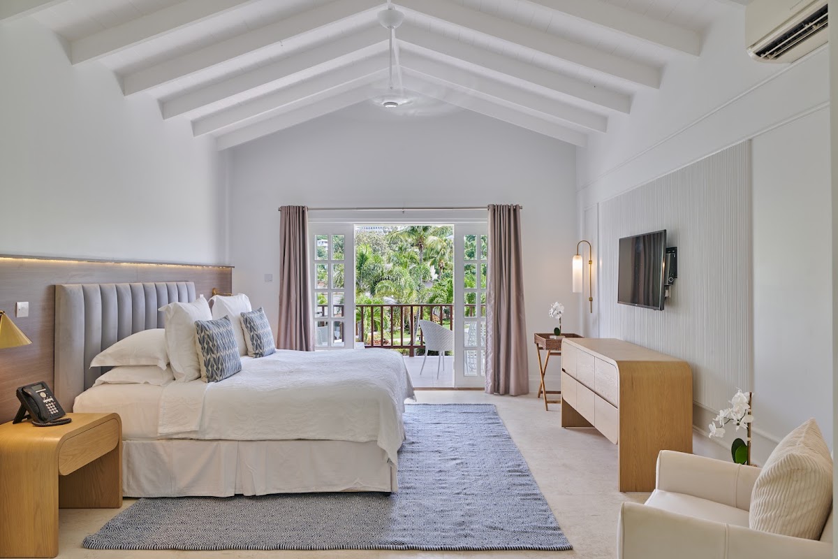 Bright bedroom with vaulted white ceiling, upholstered bed with blue pillows, and open doors leading to a balcony overlooking tropical grounds at Calabash Hotel.