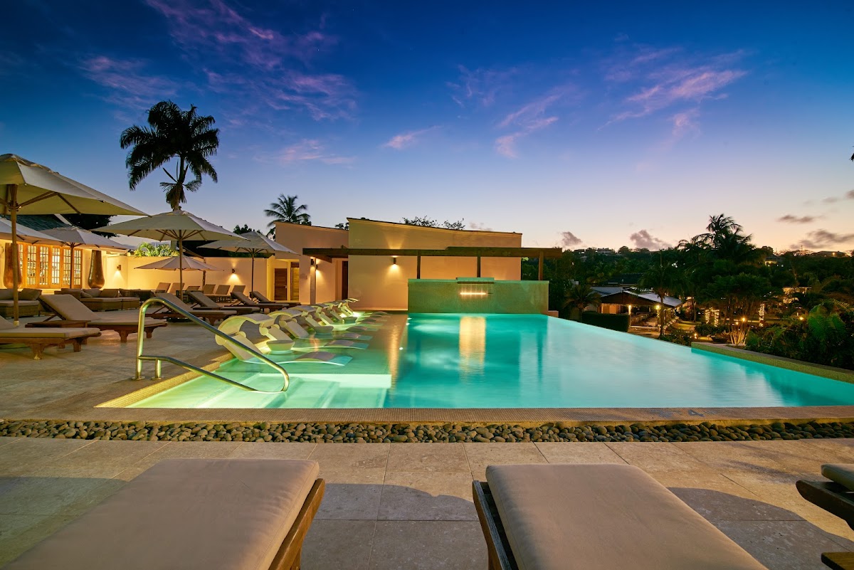 Illuminated resort pool with turquoise water, lounge chairs, umbrellas, and modern buildings at dusk in Grenada.