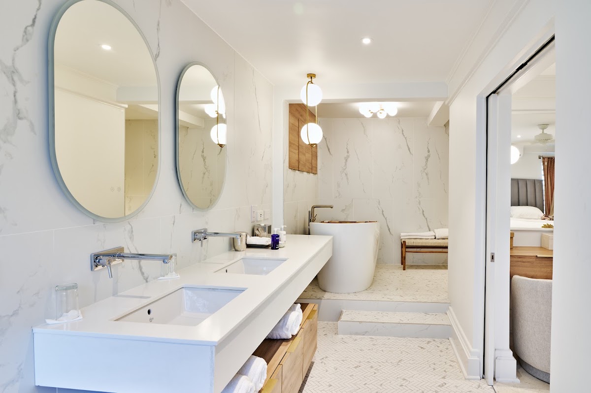 Spacious bathroom with double vanity, marble walls, two oval mirrors, freestanding soaking tub, and view into bedroom at Calabash Hotel.