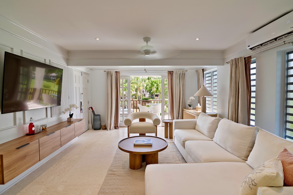 Spacious suite living room with cream seating, wooden furniture, and glass doors opening to a tropical garden view.