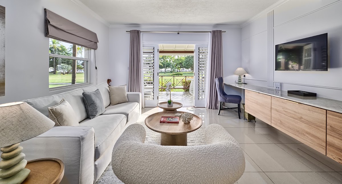 Spacious resort suite with gray sofa, curved cream chair, wooden tables, and glass doors opening to a balcony overlooking green grounds.